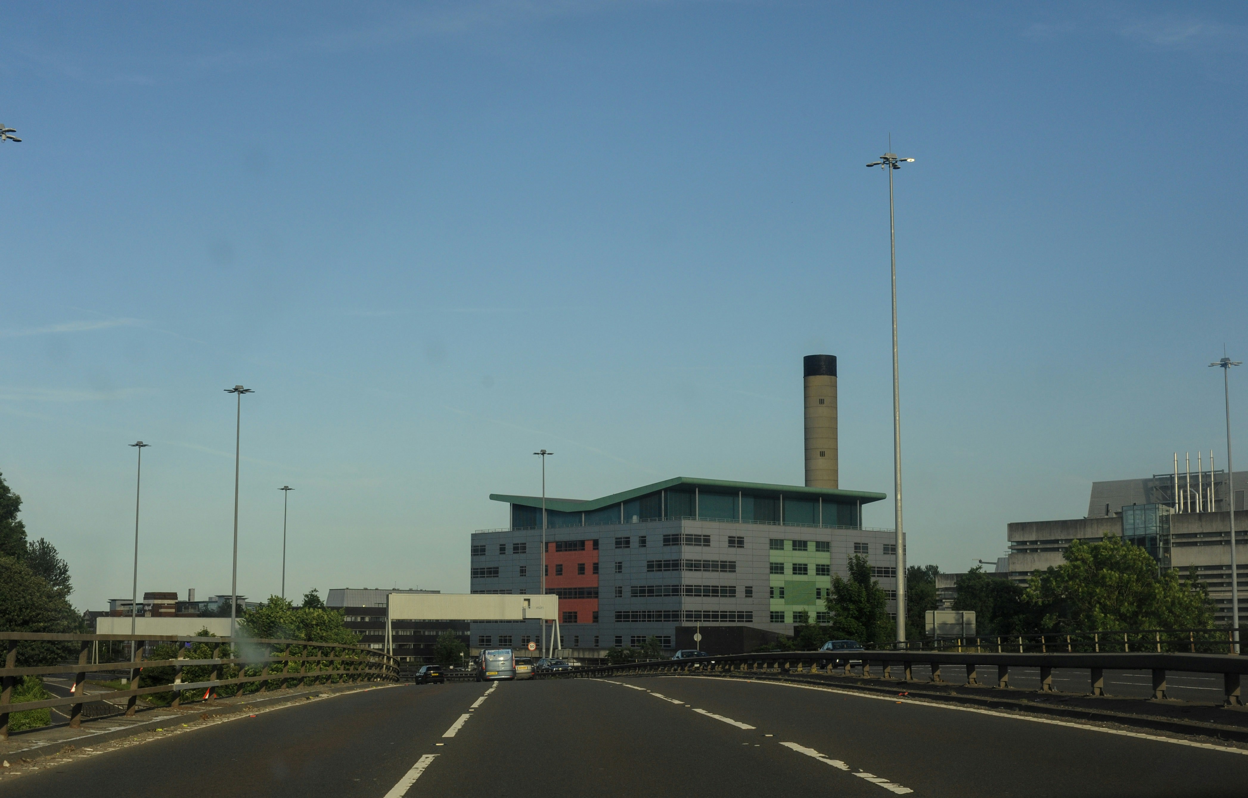 Road leading to a modern building with a tall chimney.