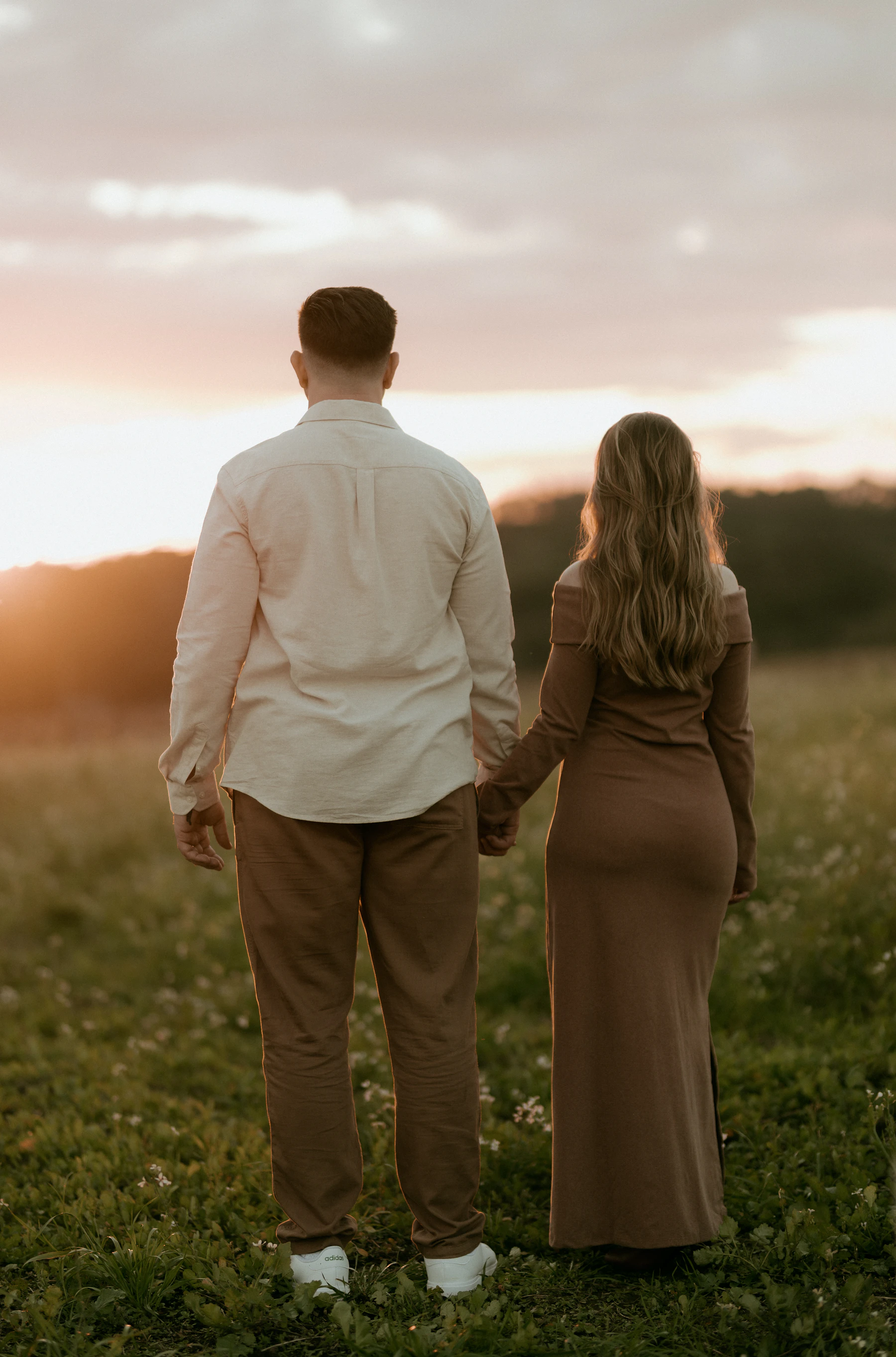 A couple walking hand in hand through a grassy field at sunset, seen from behind