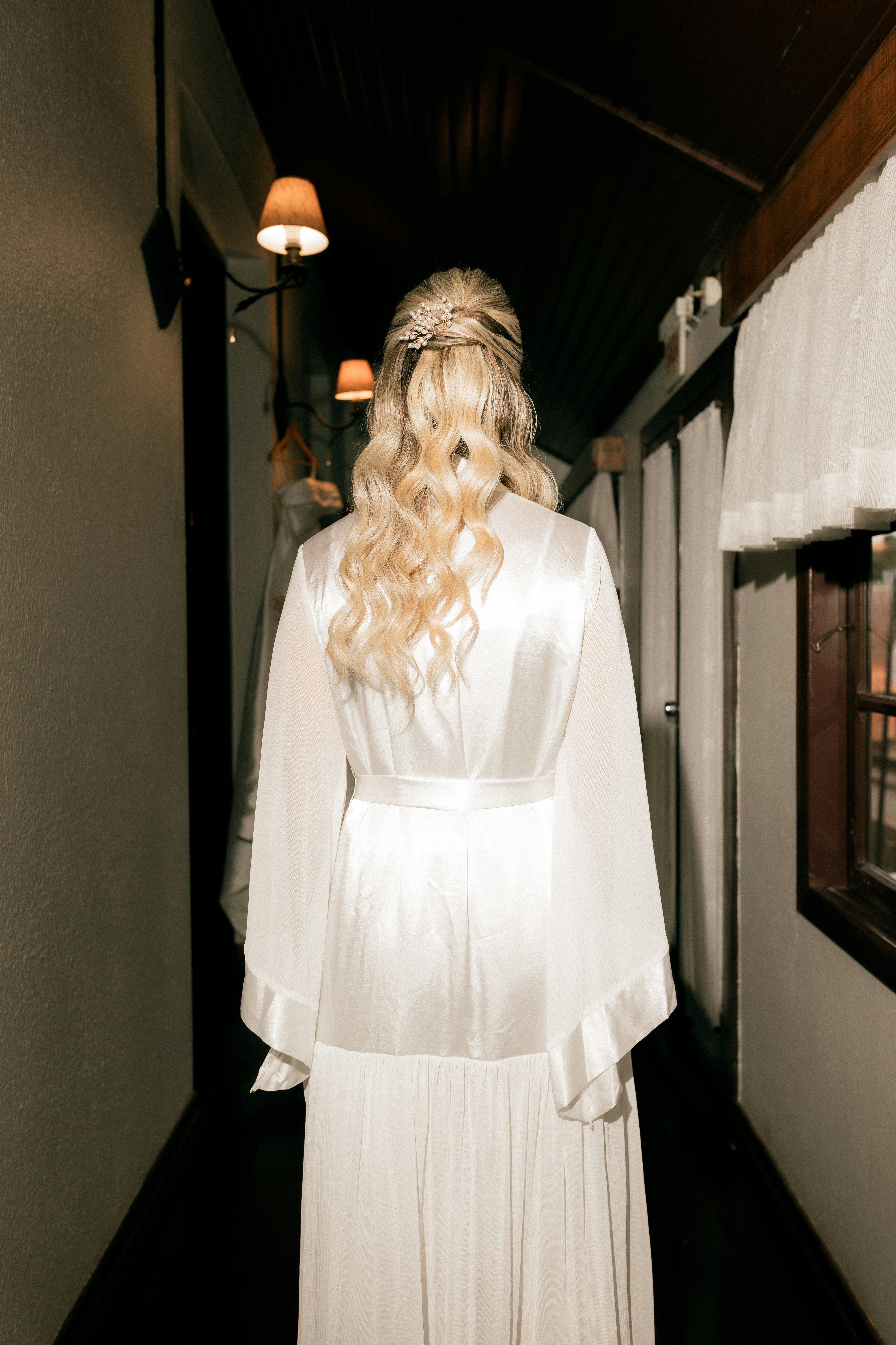 Una donna bionda in accappatoio bianco cammina lungo un corridoio buio.