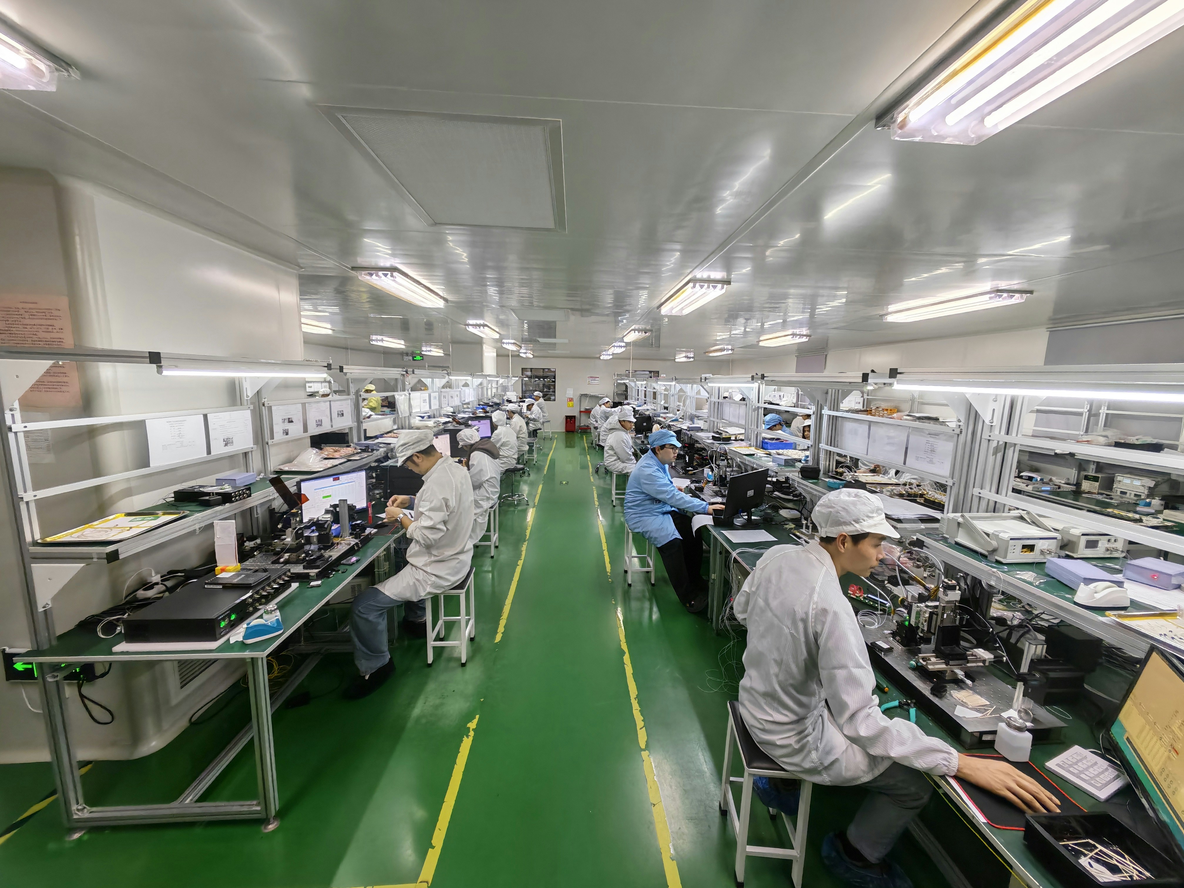 Workers in cleanroom suits assemble electronics in a long factory.