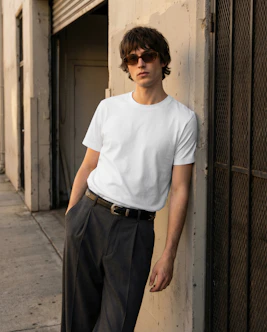 Young man in white t-shirt and sunglasses leans against wall.