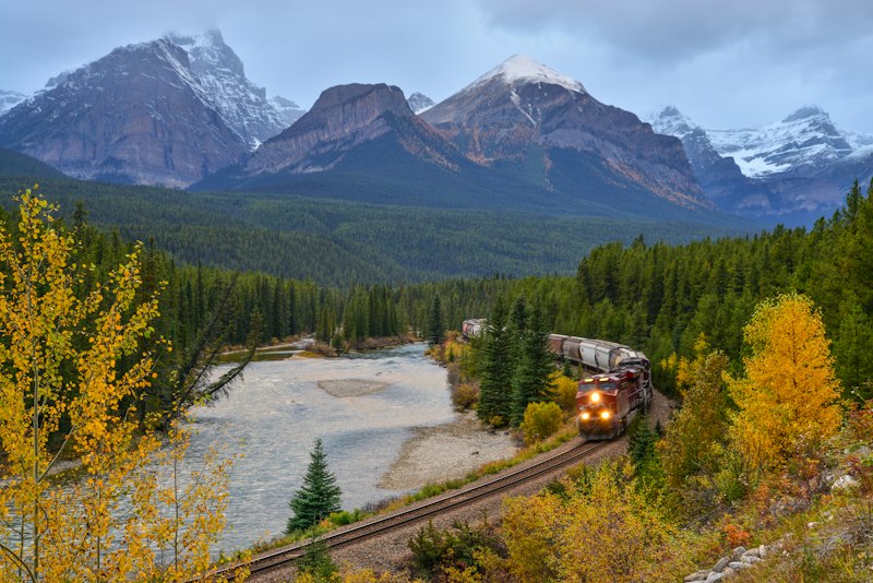 A red locomotive leading a freight train around a river bend beneath snow-dusted peaks and yellow autumn aspens