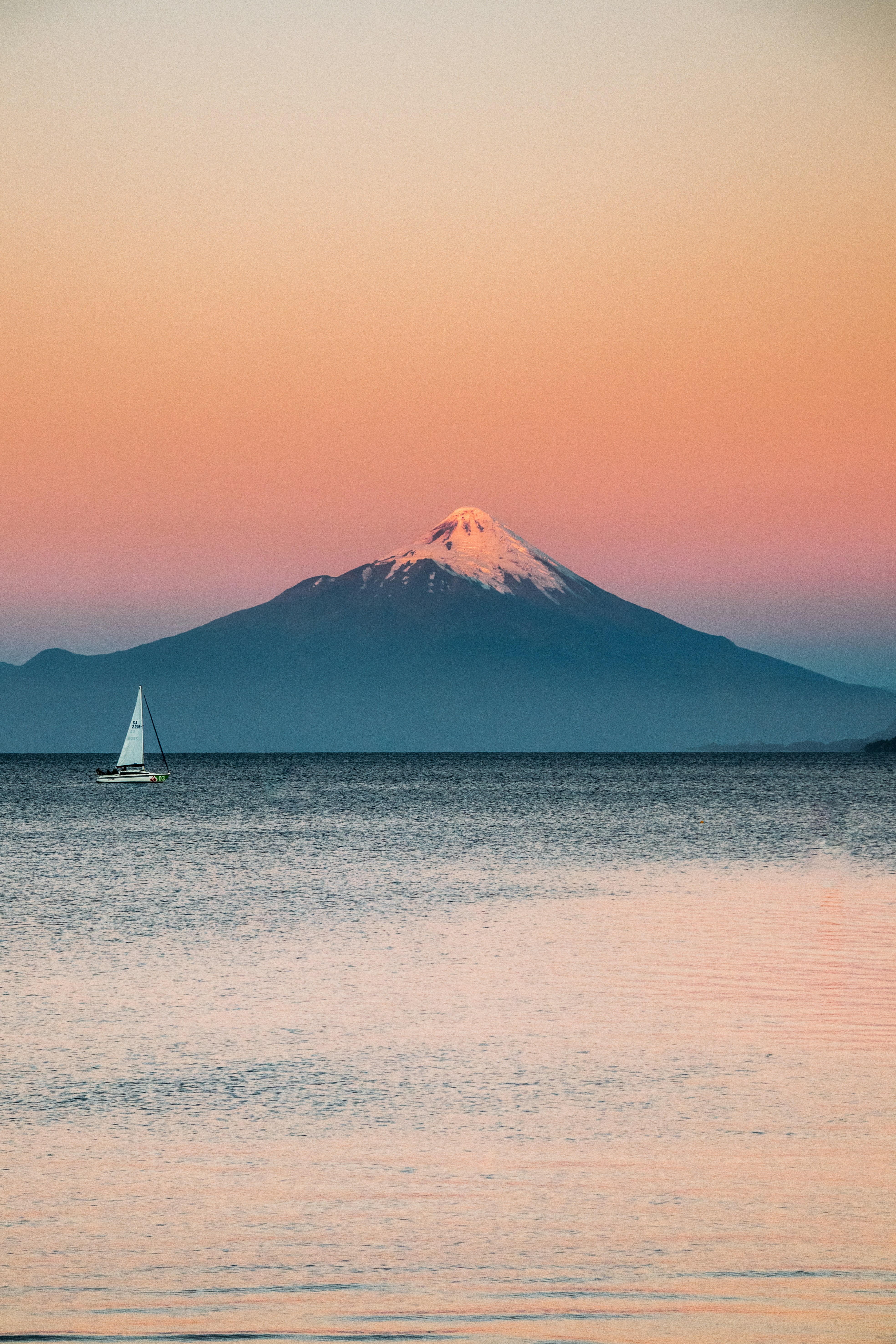 Sailboat on calm water with snow-capped mountain at sunset.