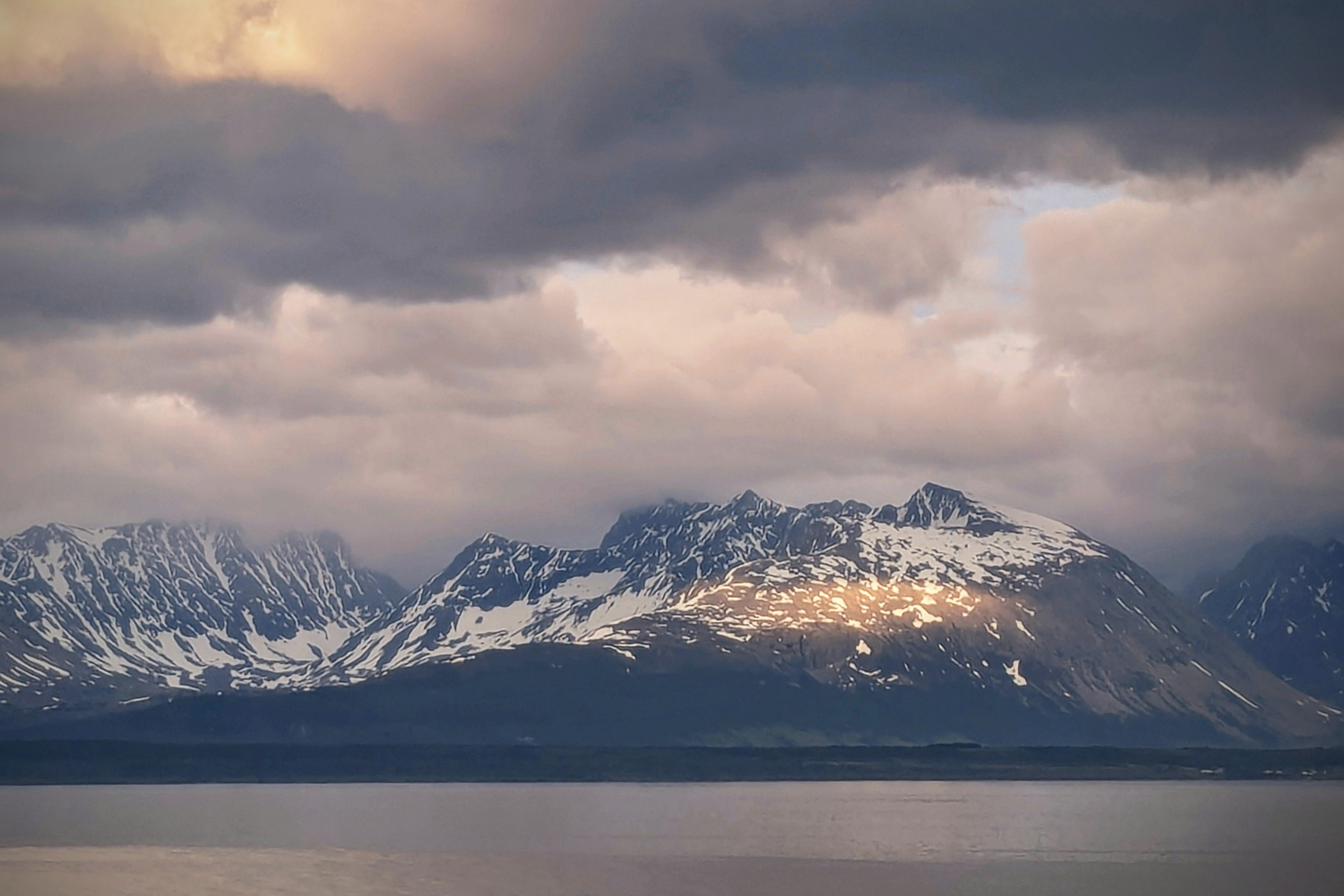 Sunlight illuminates snowy mountains above a tranquil lake under clouds.