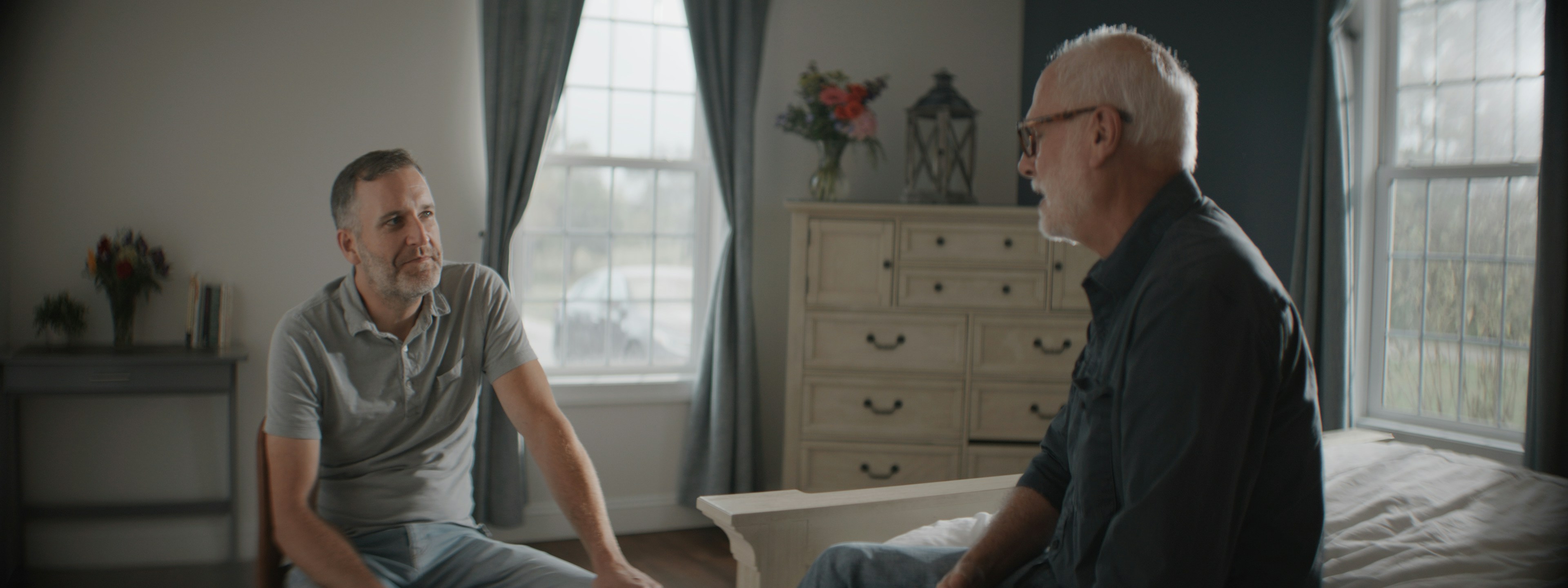 Two men in a calm, focused conversation in a sunlit bedroom, the older man leaning in as the younger listens from a chair.