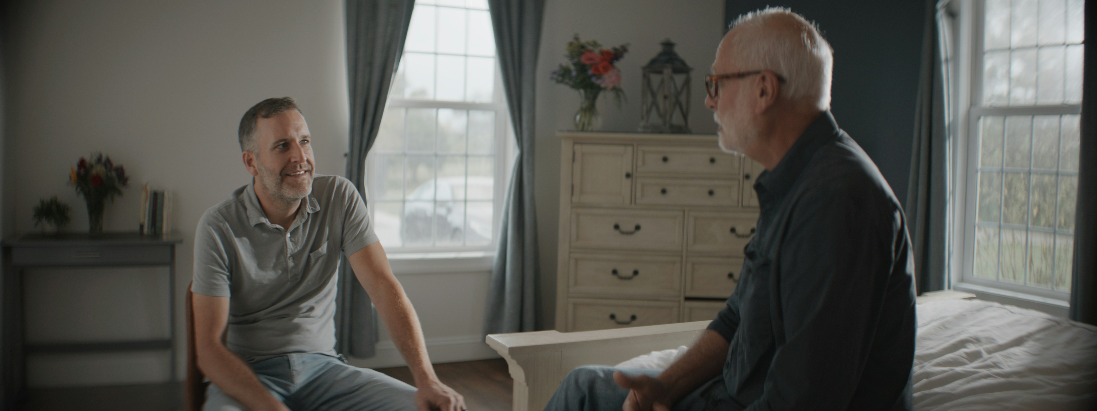 A supportive conversation between two men in a bright bedroom, the younger listening with a slight smile as the older speaks.