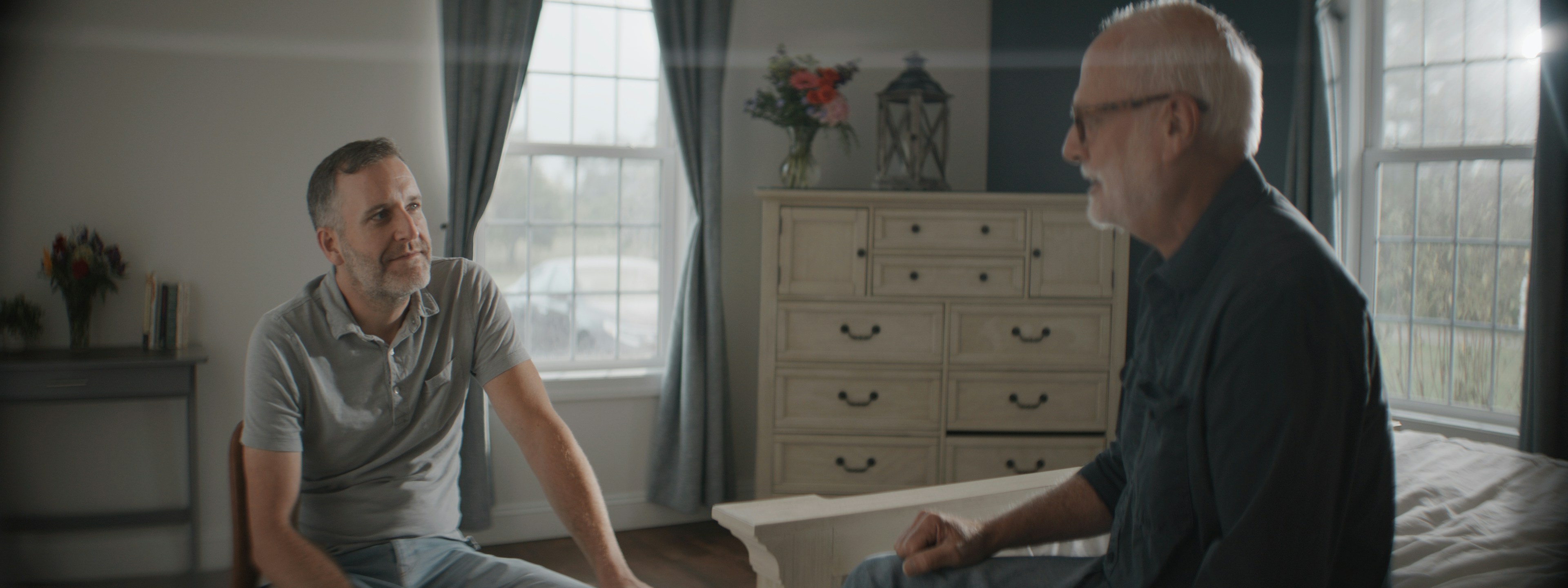 Two men conversing in a sunlit bedroom.