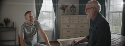 Two men conversing in a sunlit bedroom.