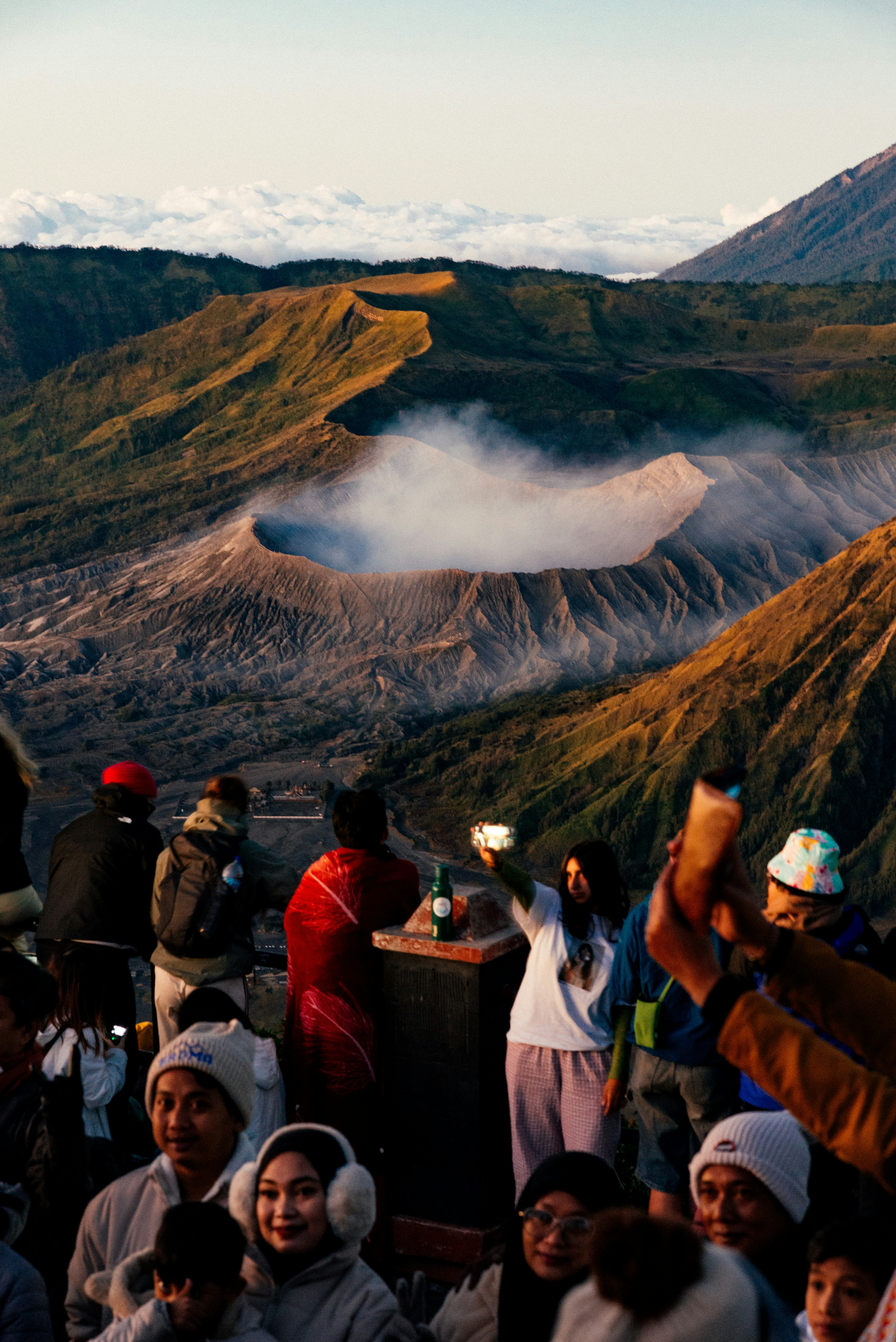 People observe a smoking volcano crater in a mountainous landscape.