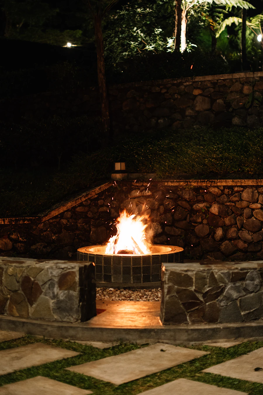 A stone fire pit glowing at night, ringed by stacked-stone walls and a paver landing
