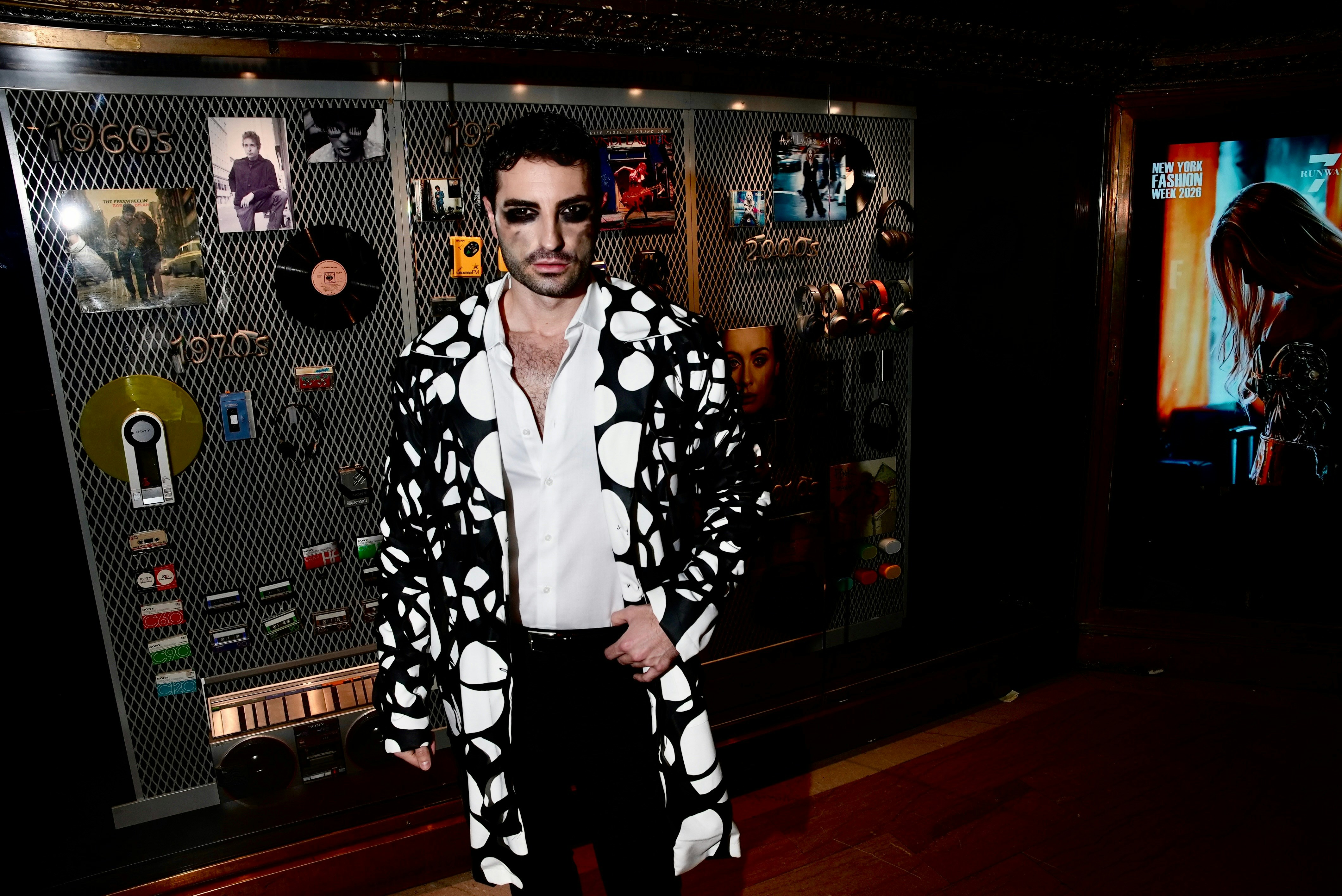 Man in patterned coat with dramatic makeup by a display.