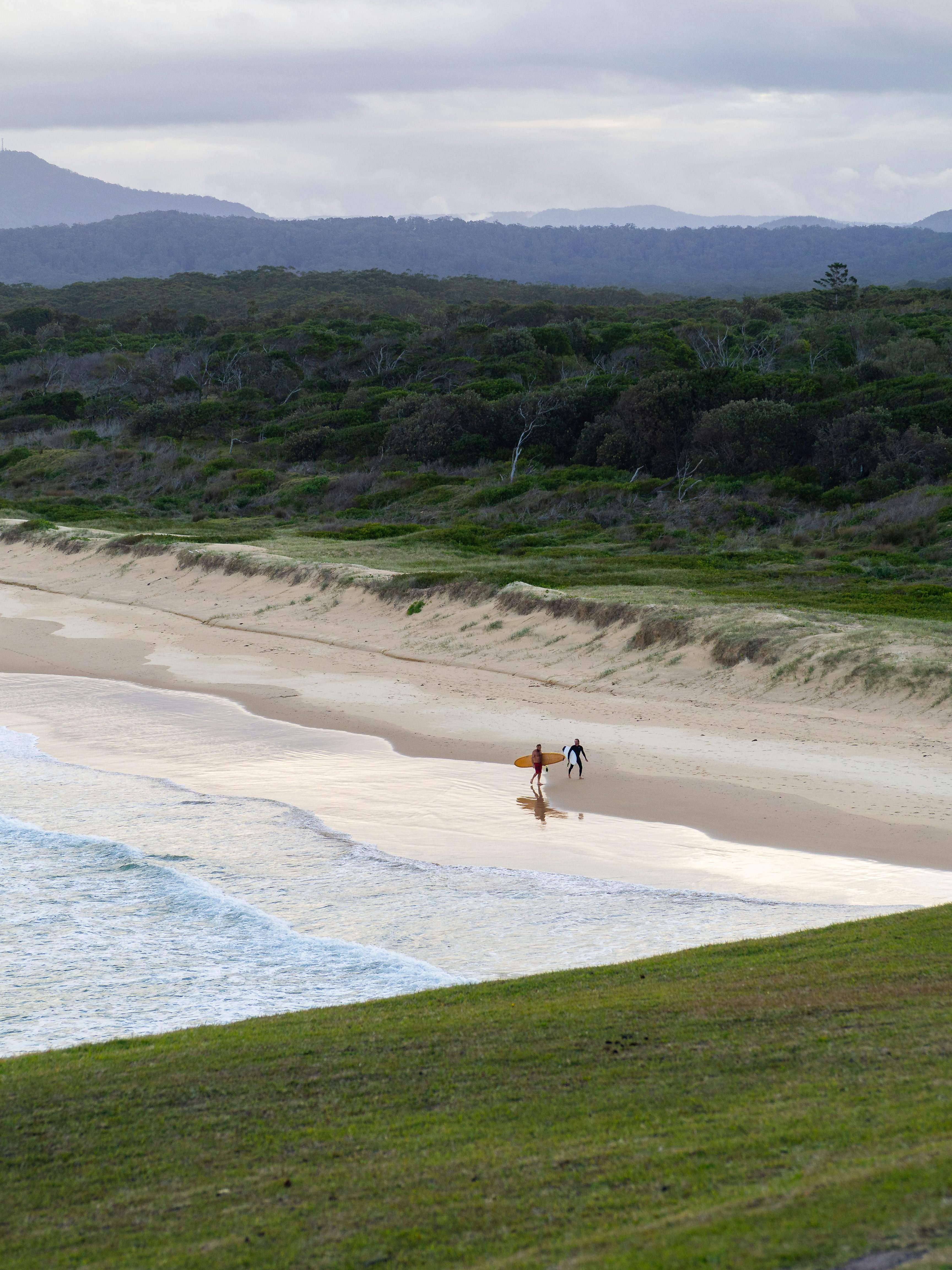 Zwei Surfer gehen an einem Sandstrand mit entfernten Bergen entlang.