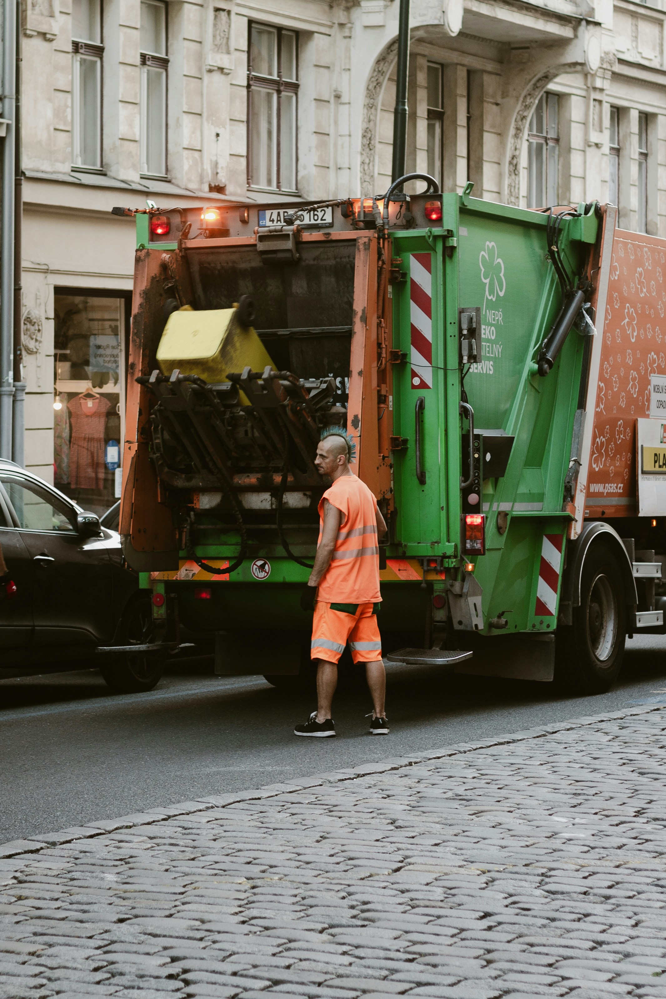 Man in orange vest stands by a green garbage truck.