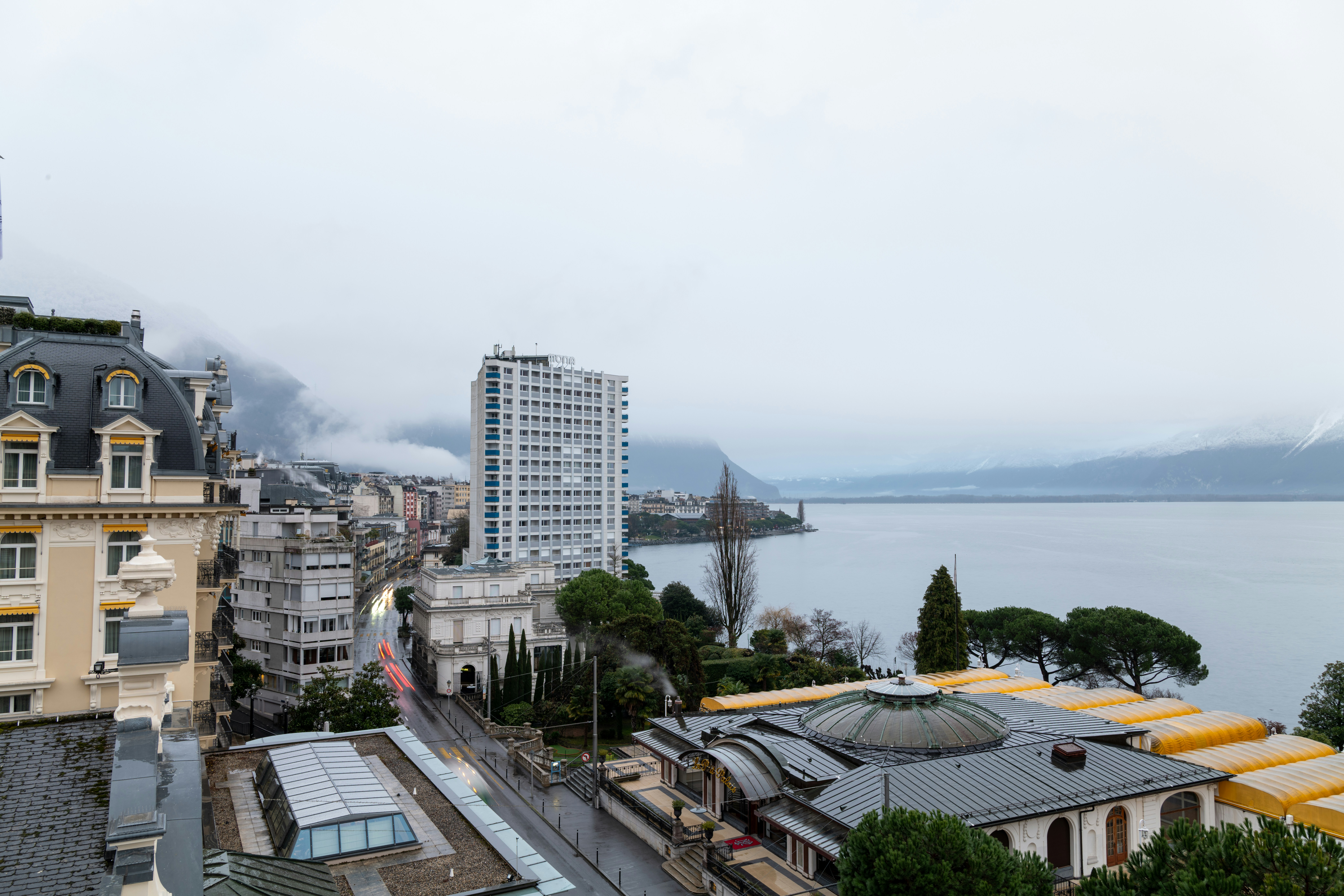 Cityscape overlooking a lake and mountains under an overcast sky.