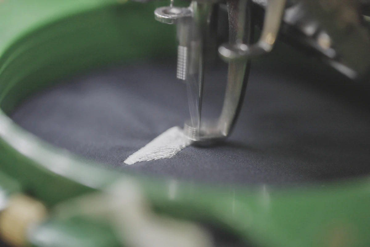 A needle laying a white satin stitch into navy cloth, held taut in a green hoop