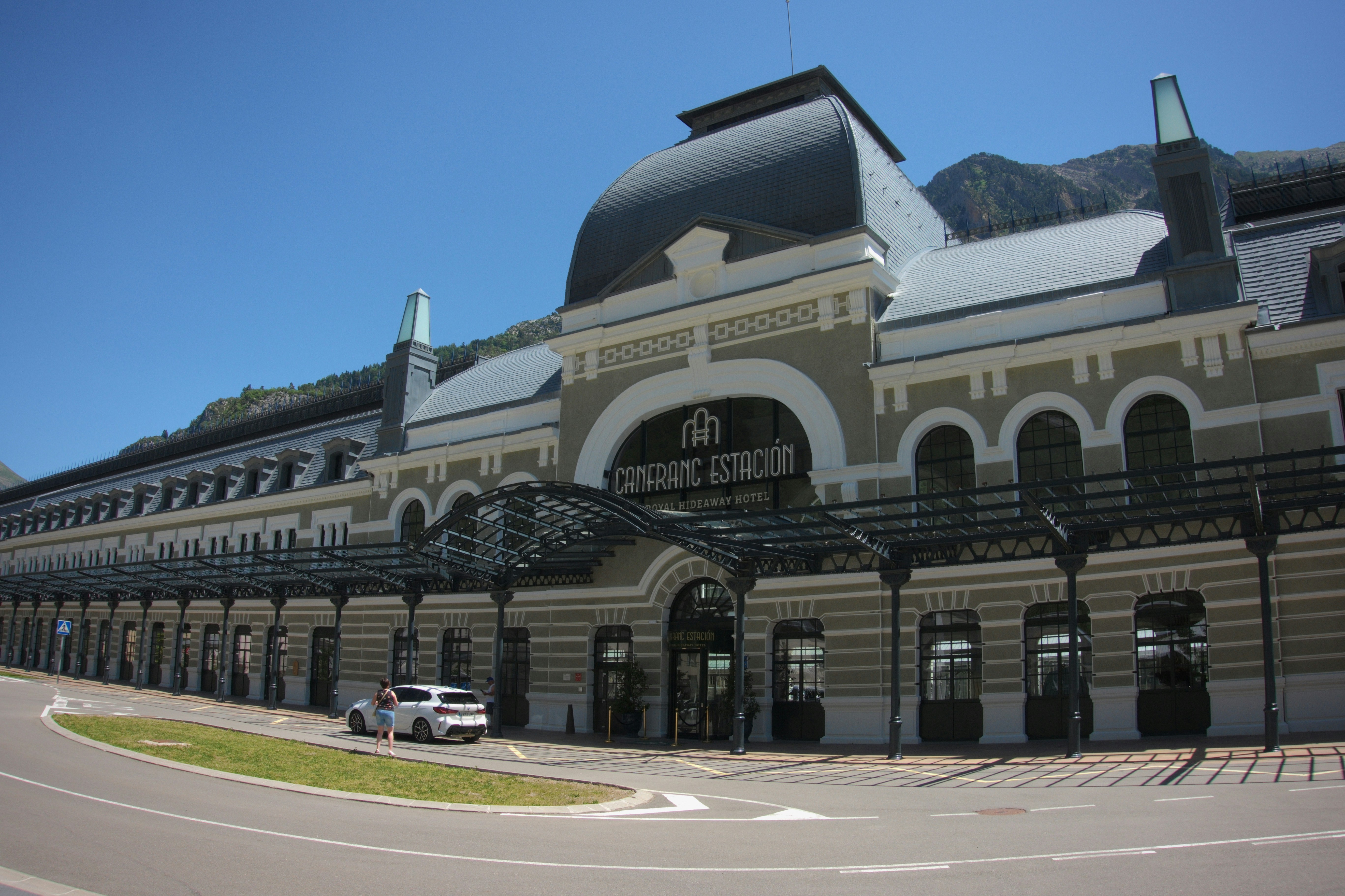 Ornate train station building with a white car and mountains.