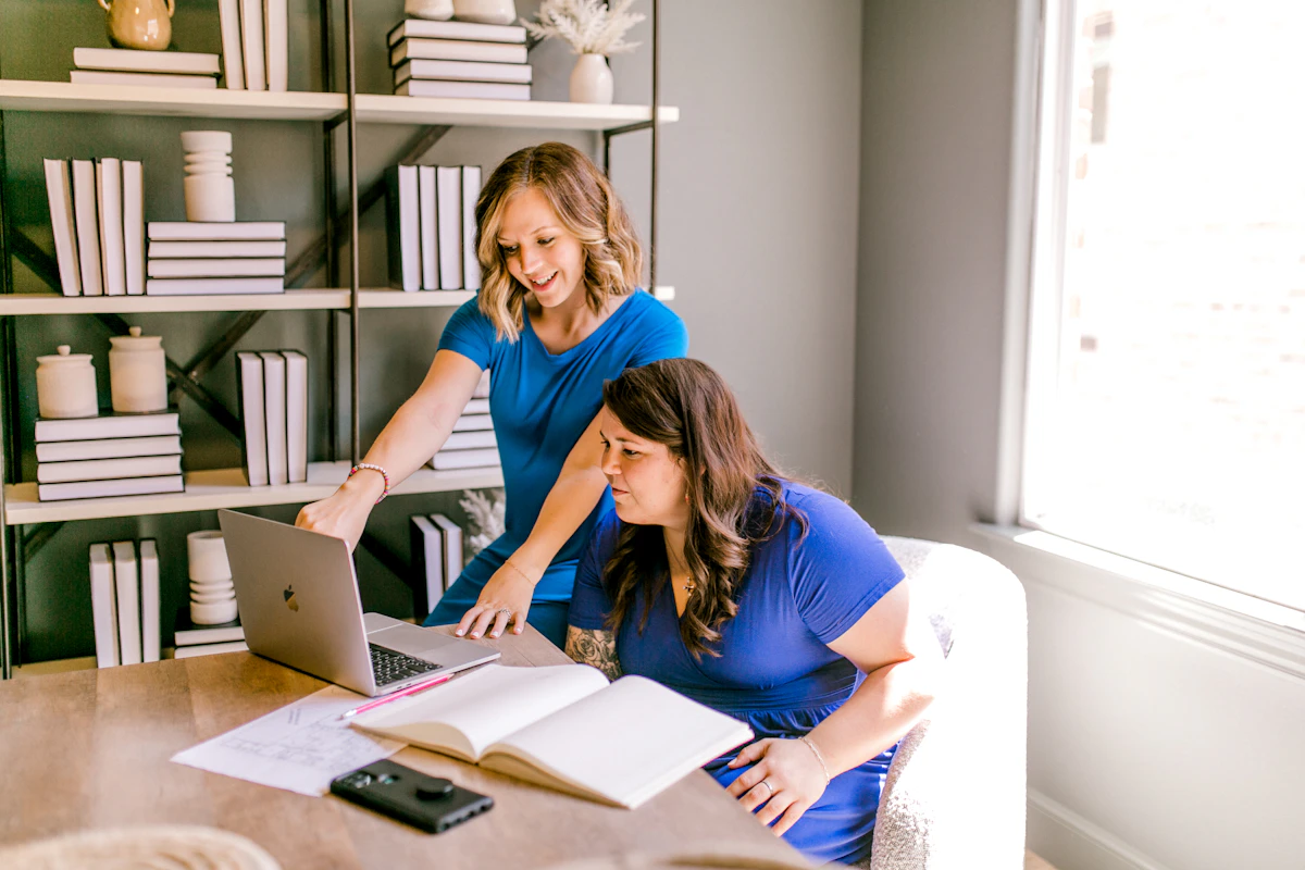 A woman leaning in to point something out on a laptop for a seated colleague by a bright window
