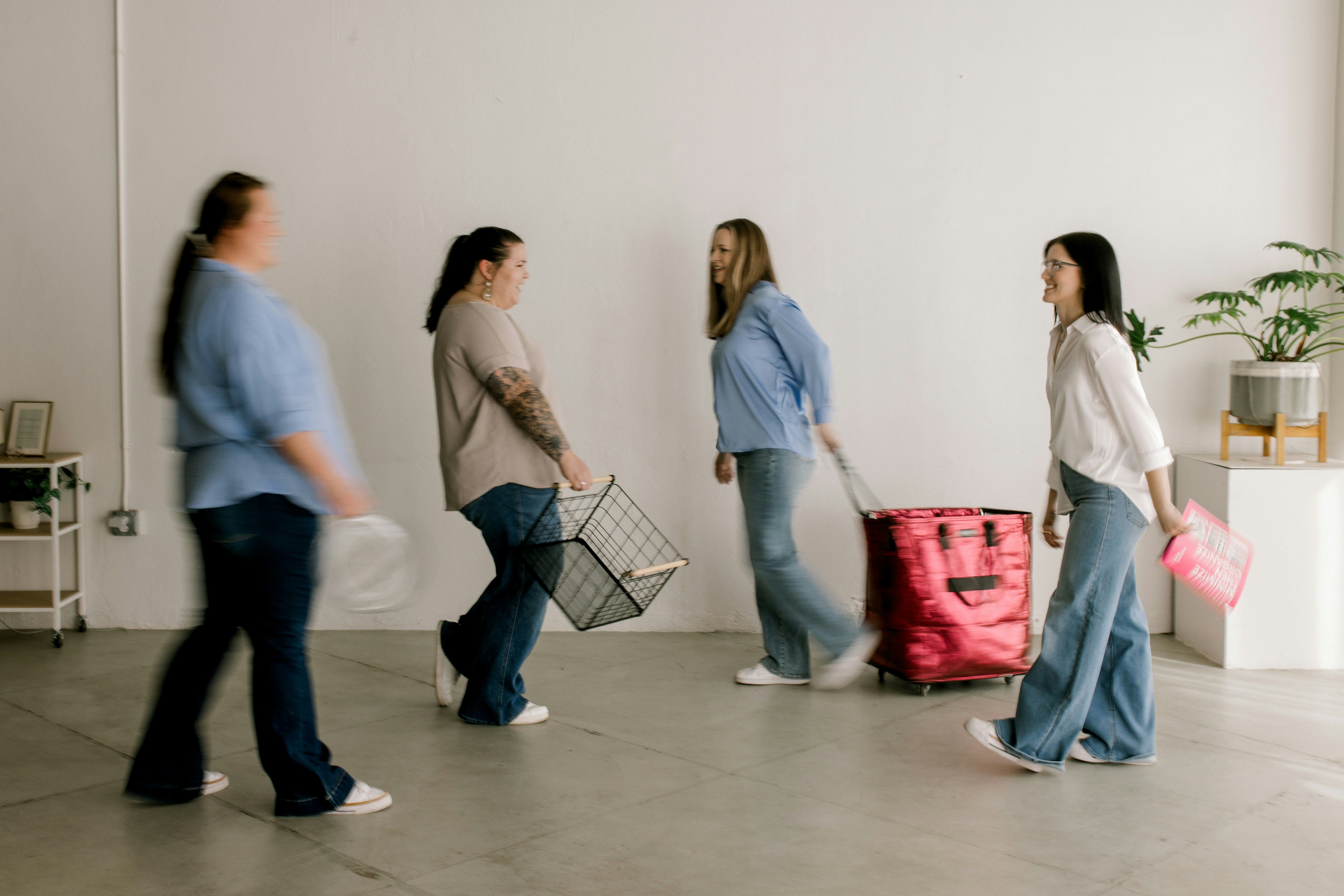 Four women walk across a minimalist white room with belongings.