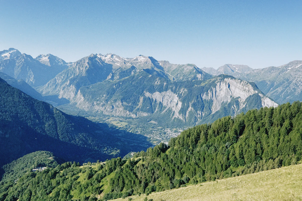 Forested green slopes falling to a valley town, with grey rock peaks and snow patches behind
