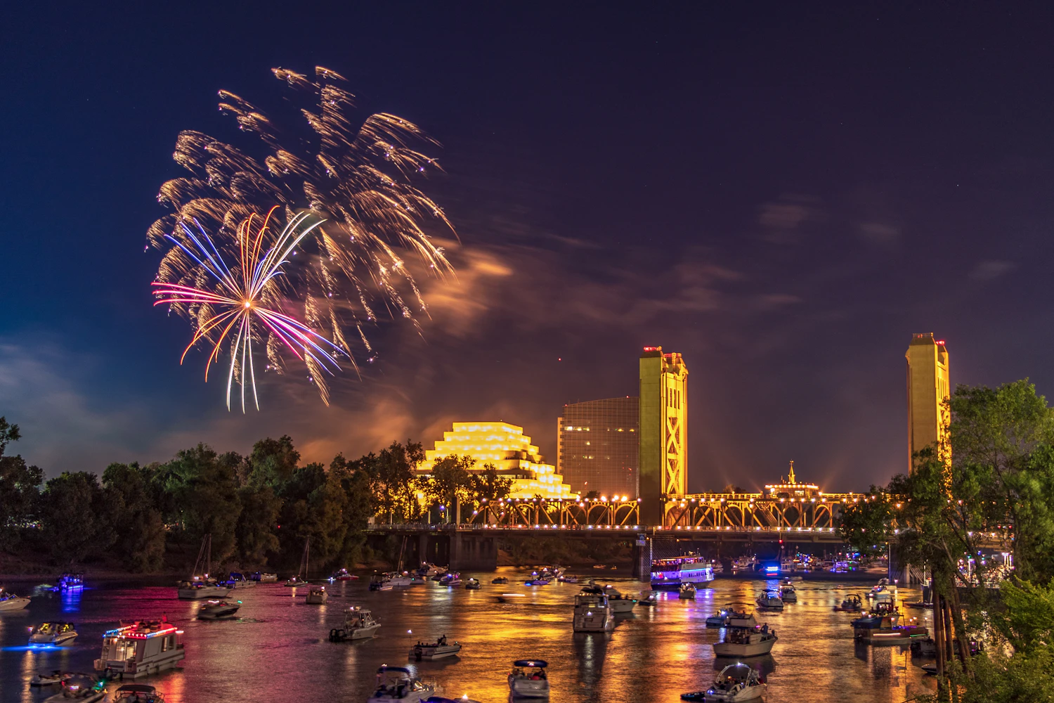 Fireworks reflected in a river double the display and tie the bursts to a recognisable skyline, something a plain black sky cannot do.