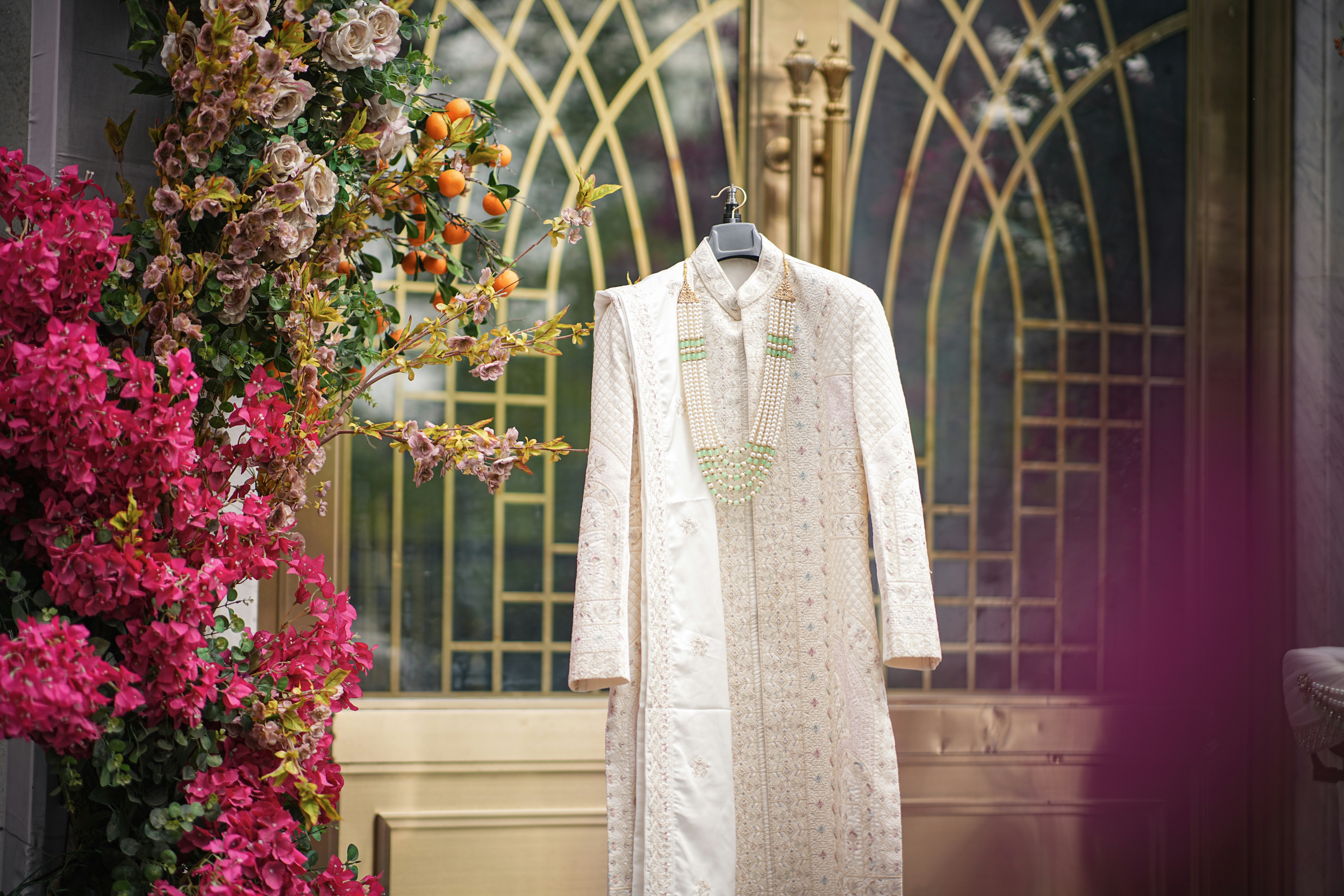 Cream sherwani hangs on ornate golden door with vibrant flowers.