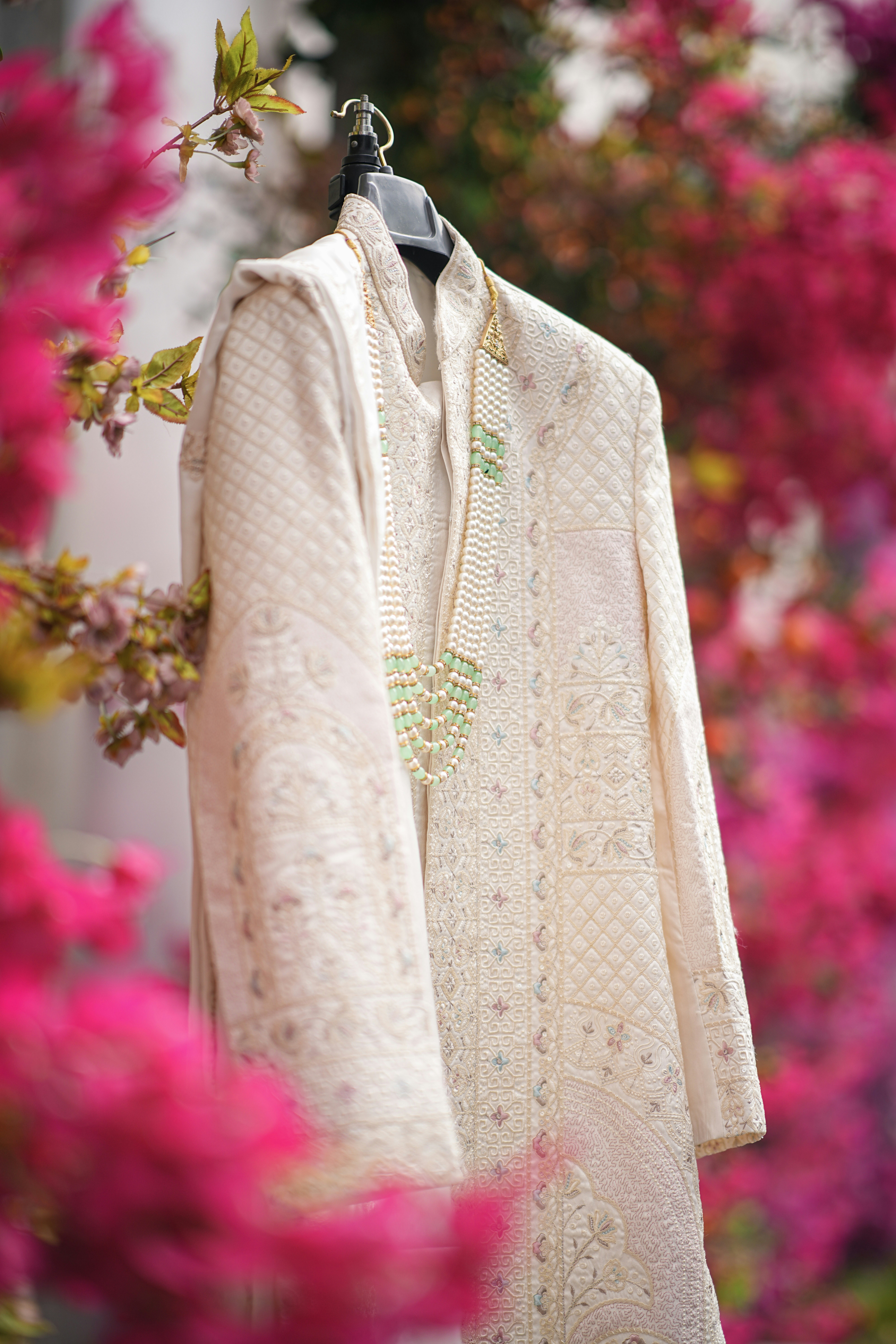 A cream sherwani with a necklace hangs among pink flowers.