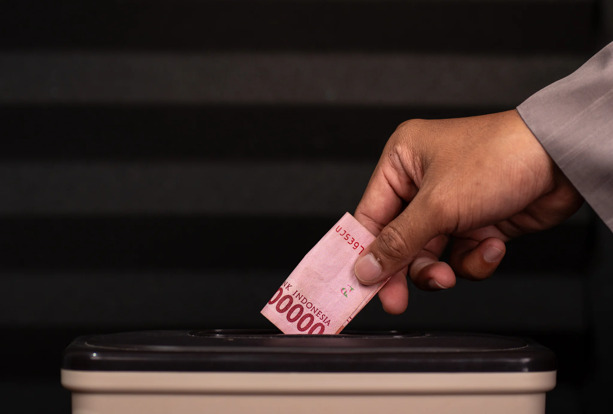 A hand placing a folded banknote into the slot of a donation box