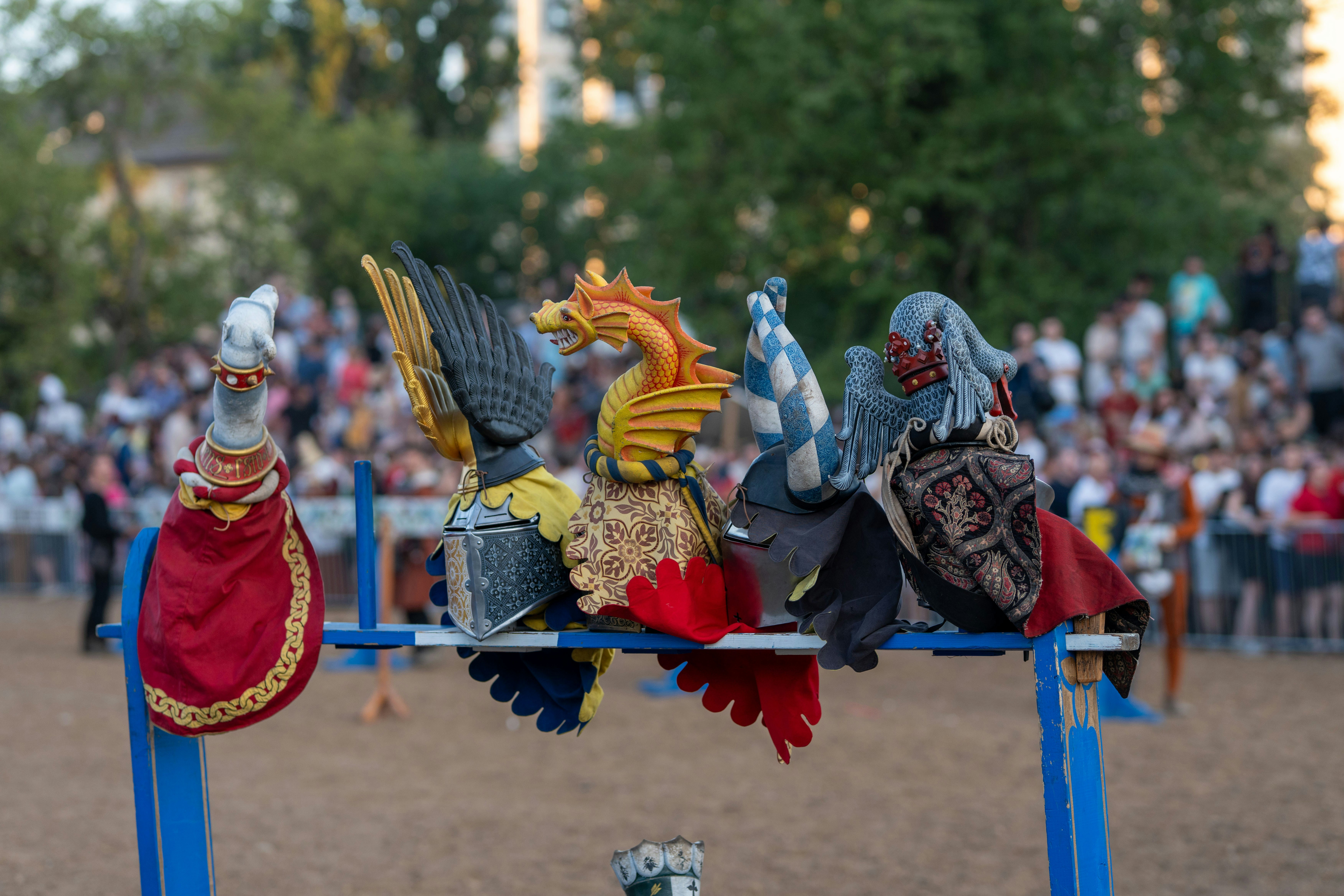 Five ornate medieval helmets with colorful crests on display.