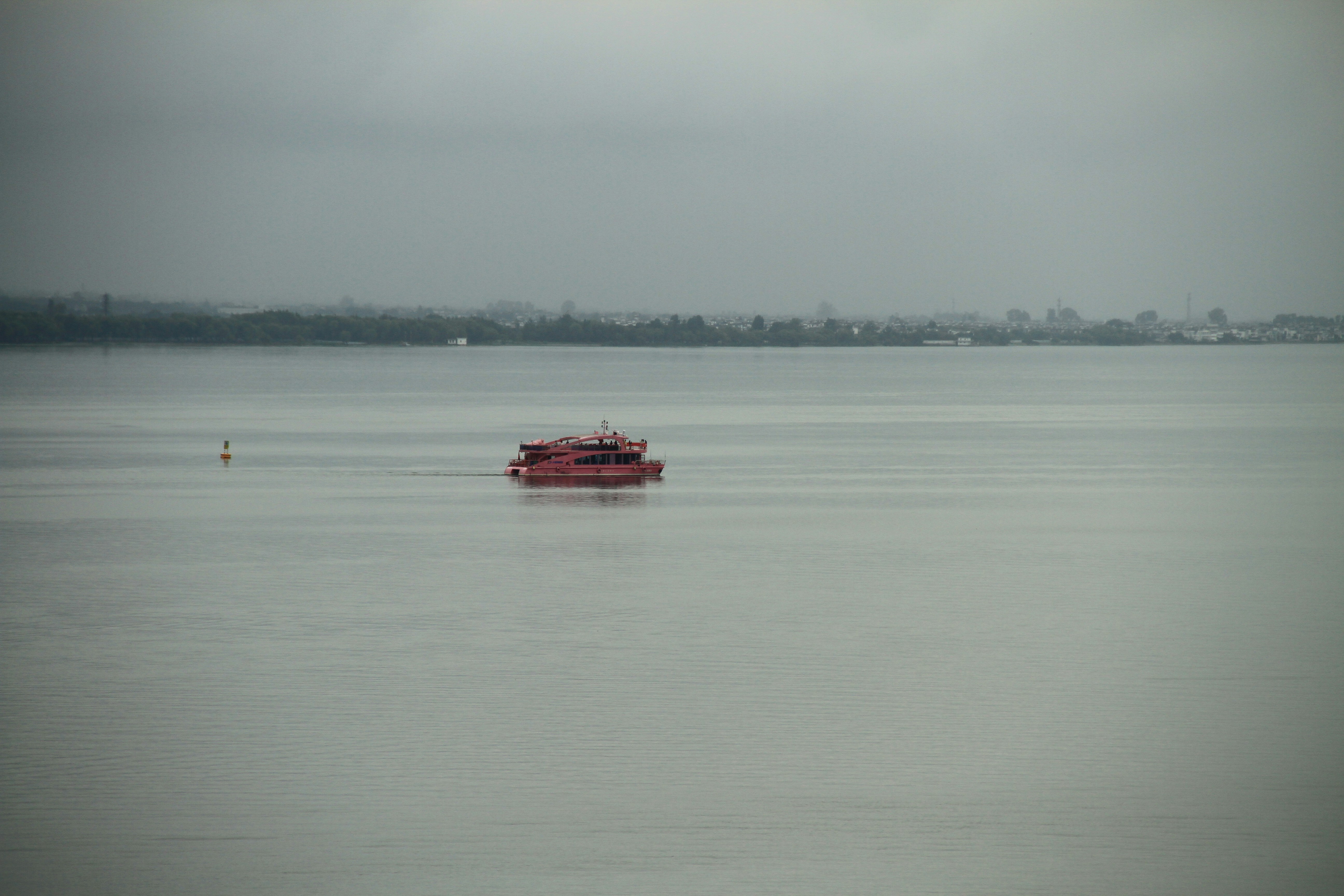 Red ferry boat crosses calm grey water under overcast sky.