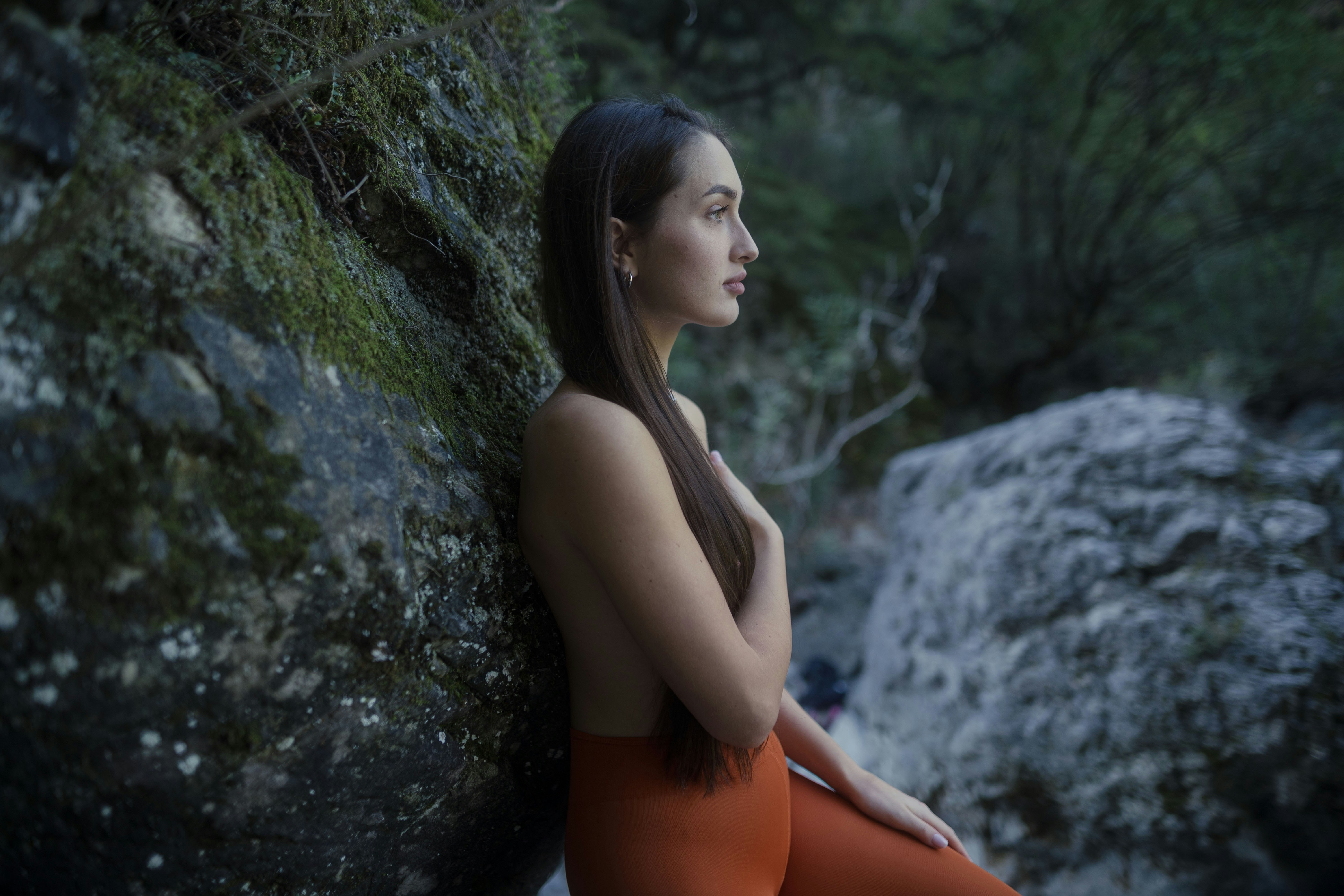 Mujer con pantalones naranjas apoyada en una roca musgosa en la naturaleza.