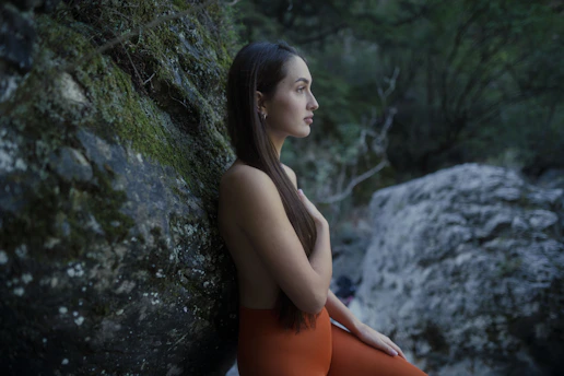 Woman in orange pants leans on mossy rock in nature.