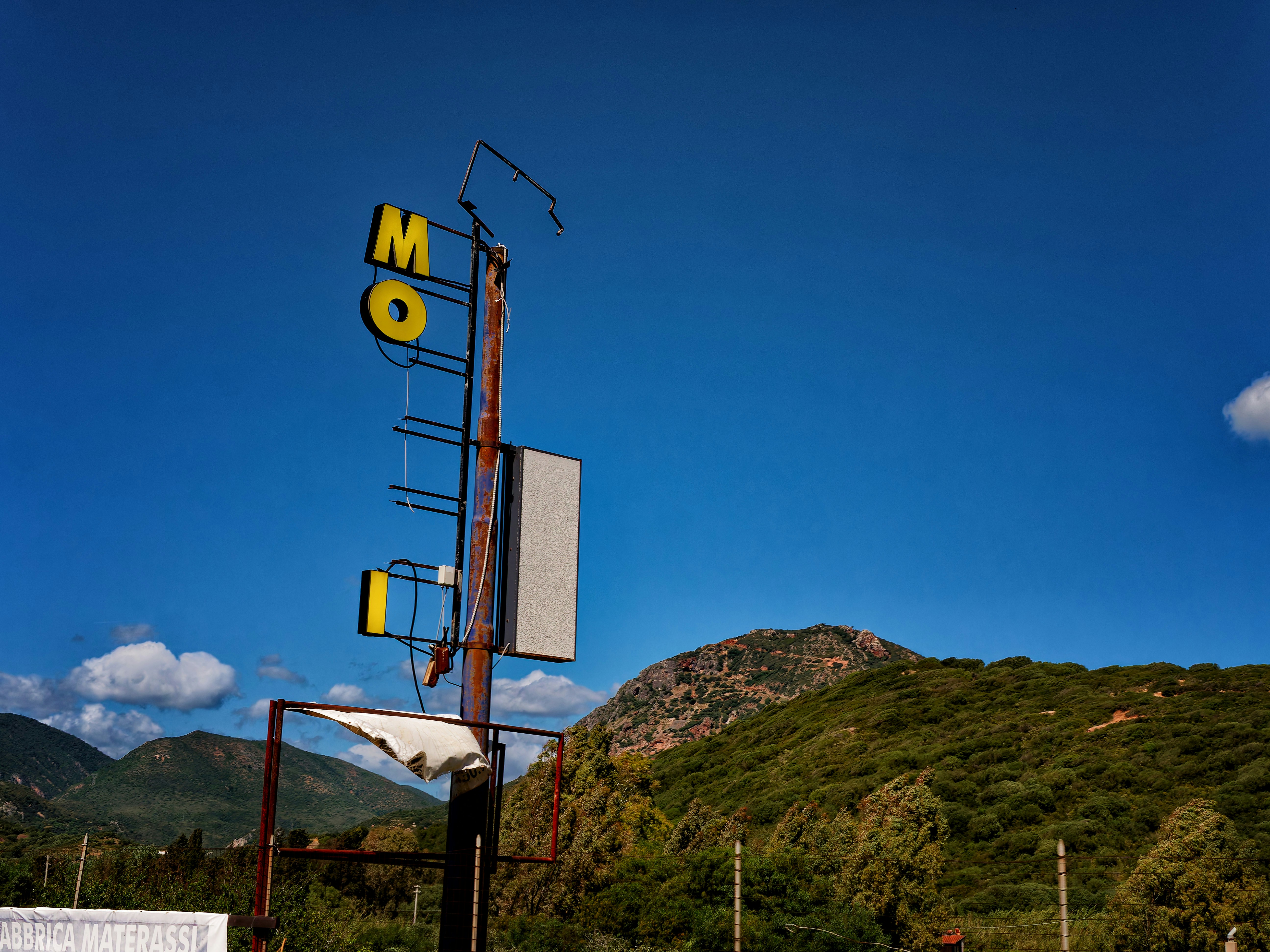 Weathered "mo" sign against blue sky and green hills.
