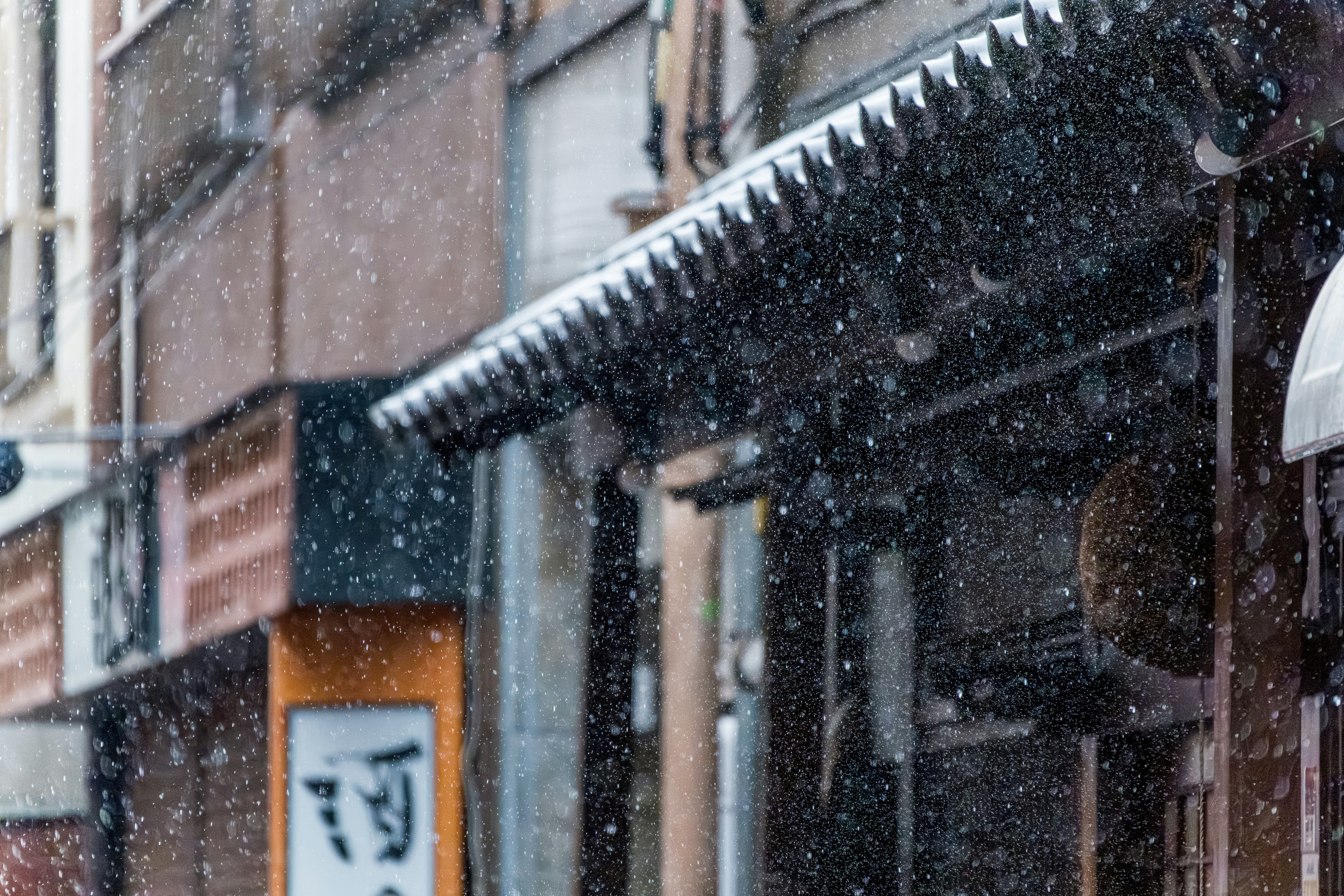 Snow falls on a street with buildings and an awning.