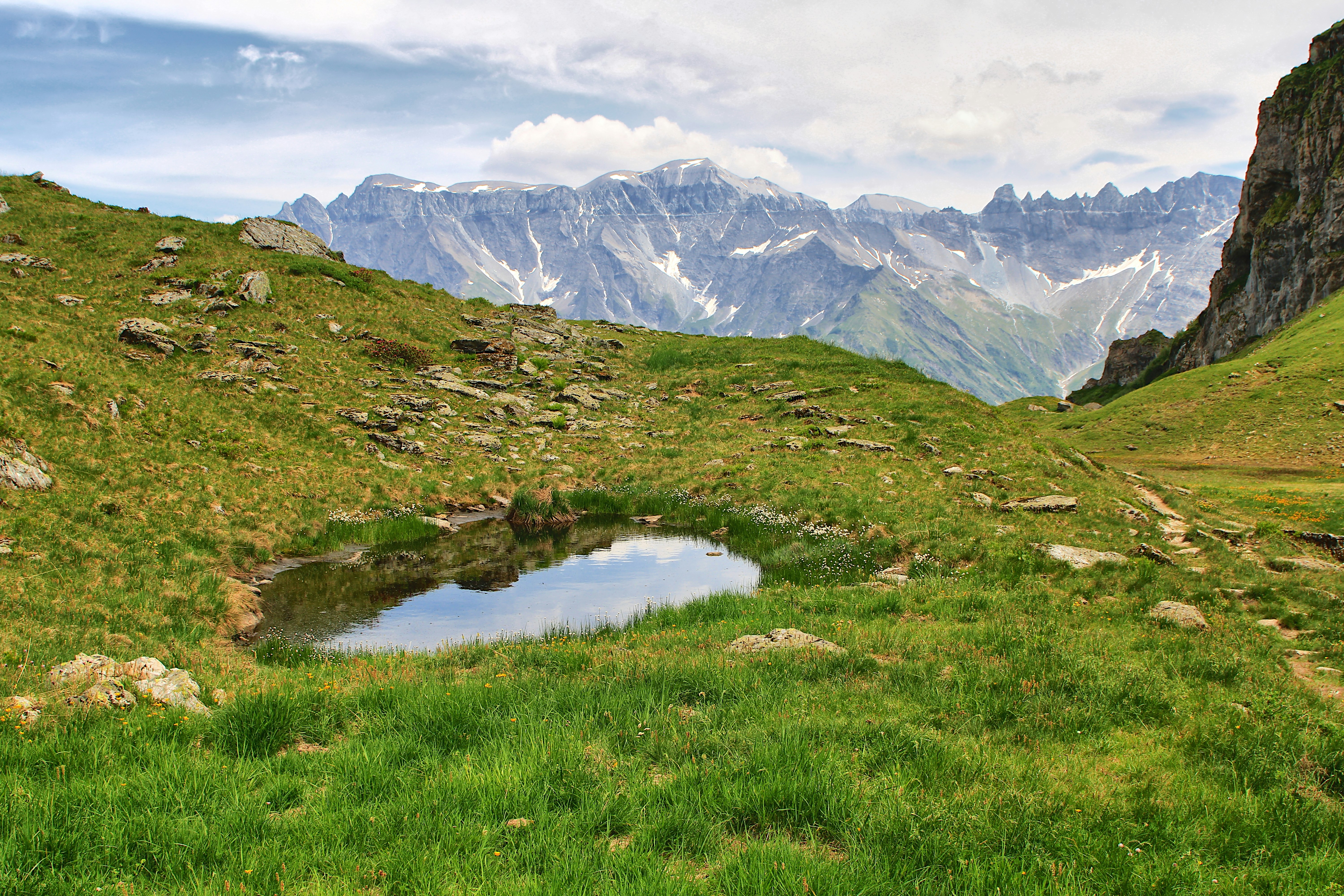 El estanque alpino refleja montañas nevadas entre colinas verdes.