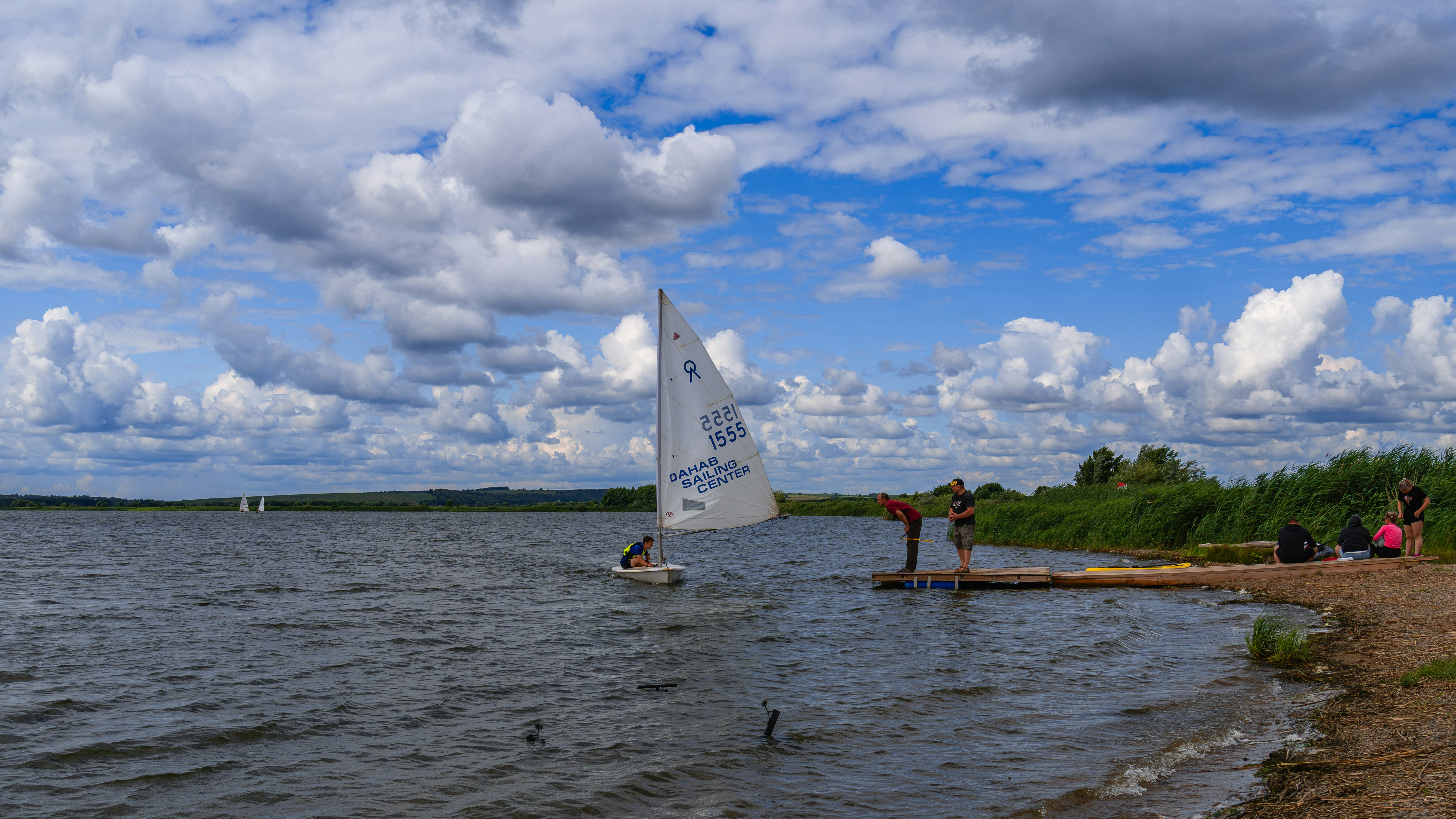 Kleines Segelboot, das sich einem Seeufer mit Menschen und Wolken nähert.