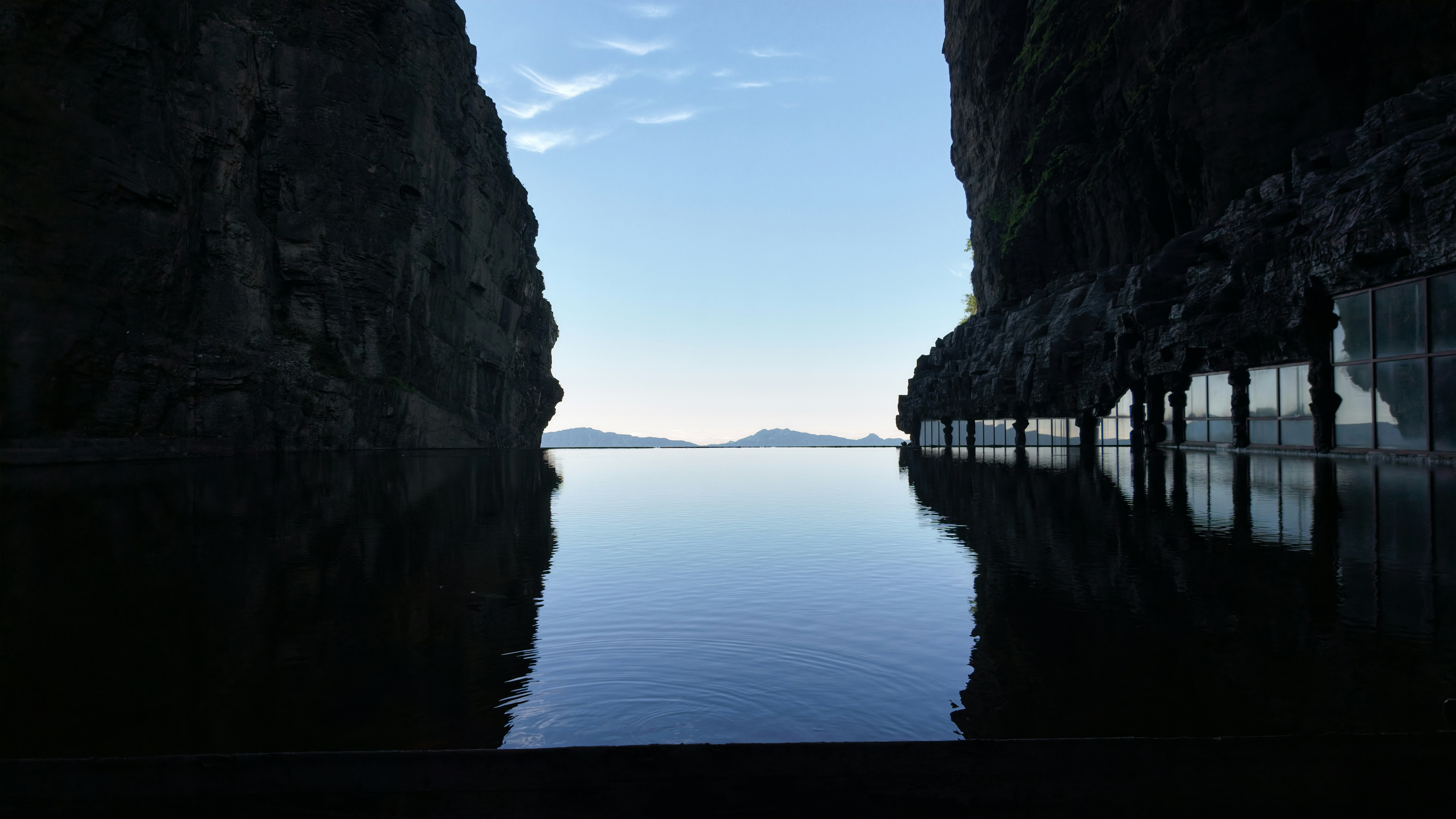 A tranquil water passage between towering cliff faces leads the eye toward a distant ridge across a calm, reflective sea. The scene emphasizes symmetry and the stillness of the environment; a sign of a coastal landscape or architectural waterway.