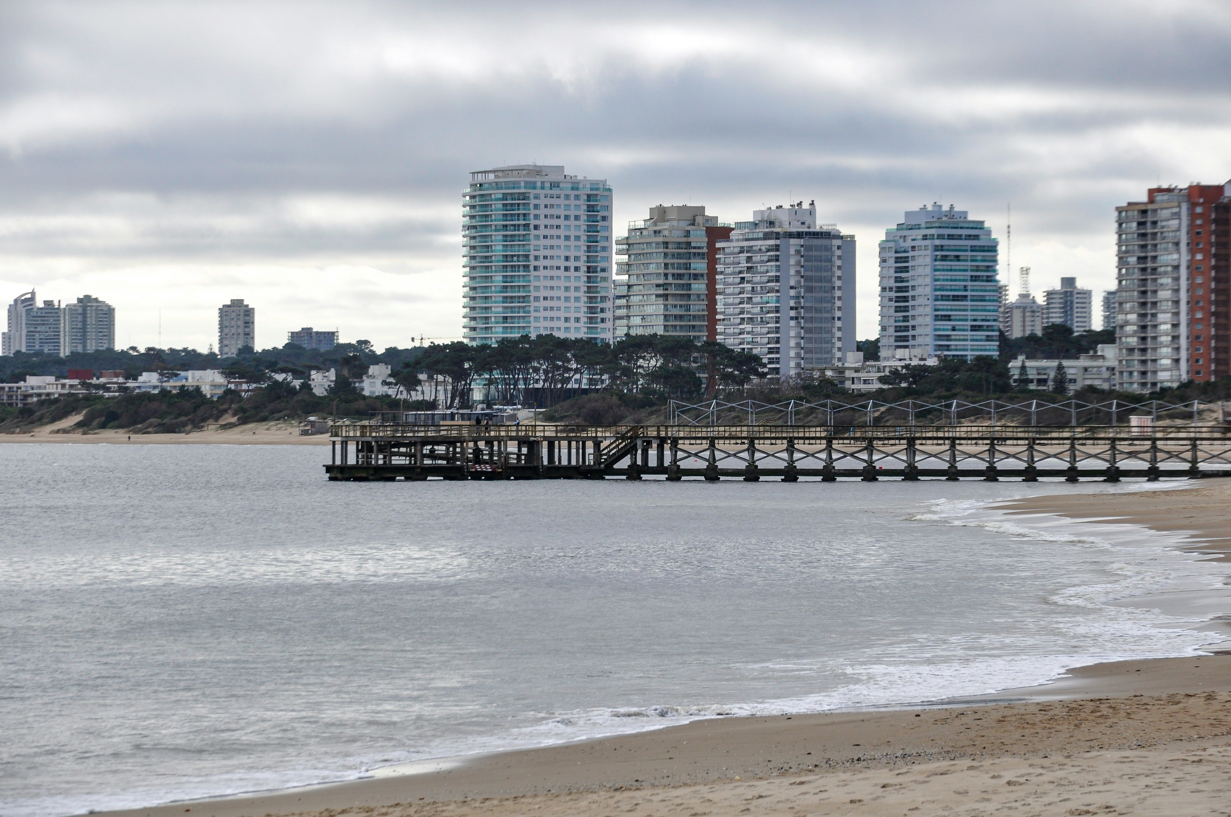 City skyline, pier, and sandy beach under a cloudy sky.