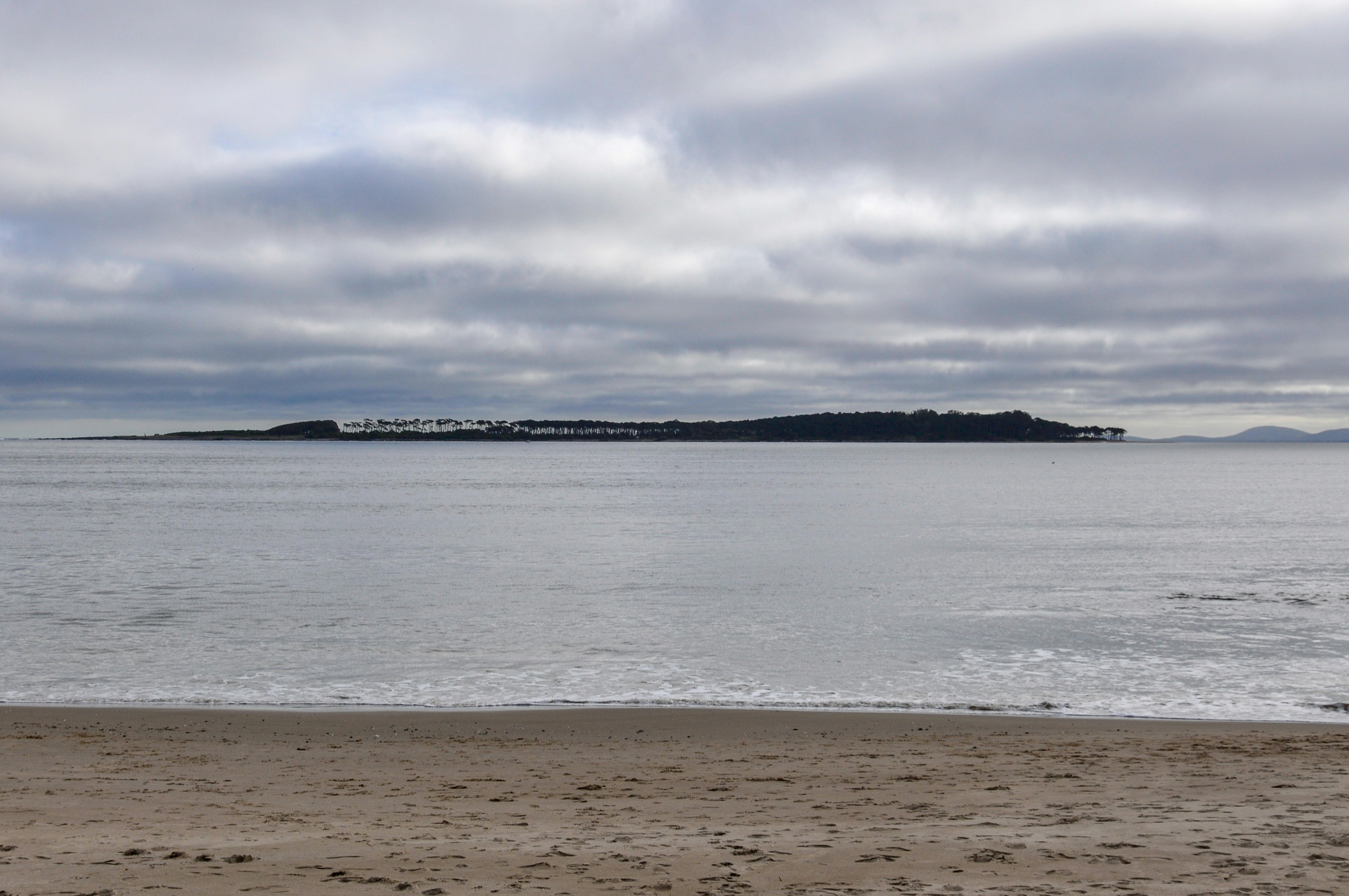 Beach, calm ocean, and distant island under cloudy sky.