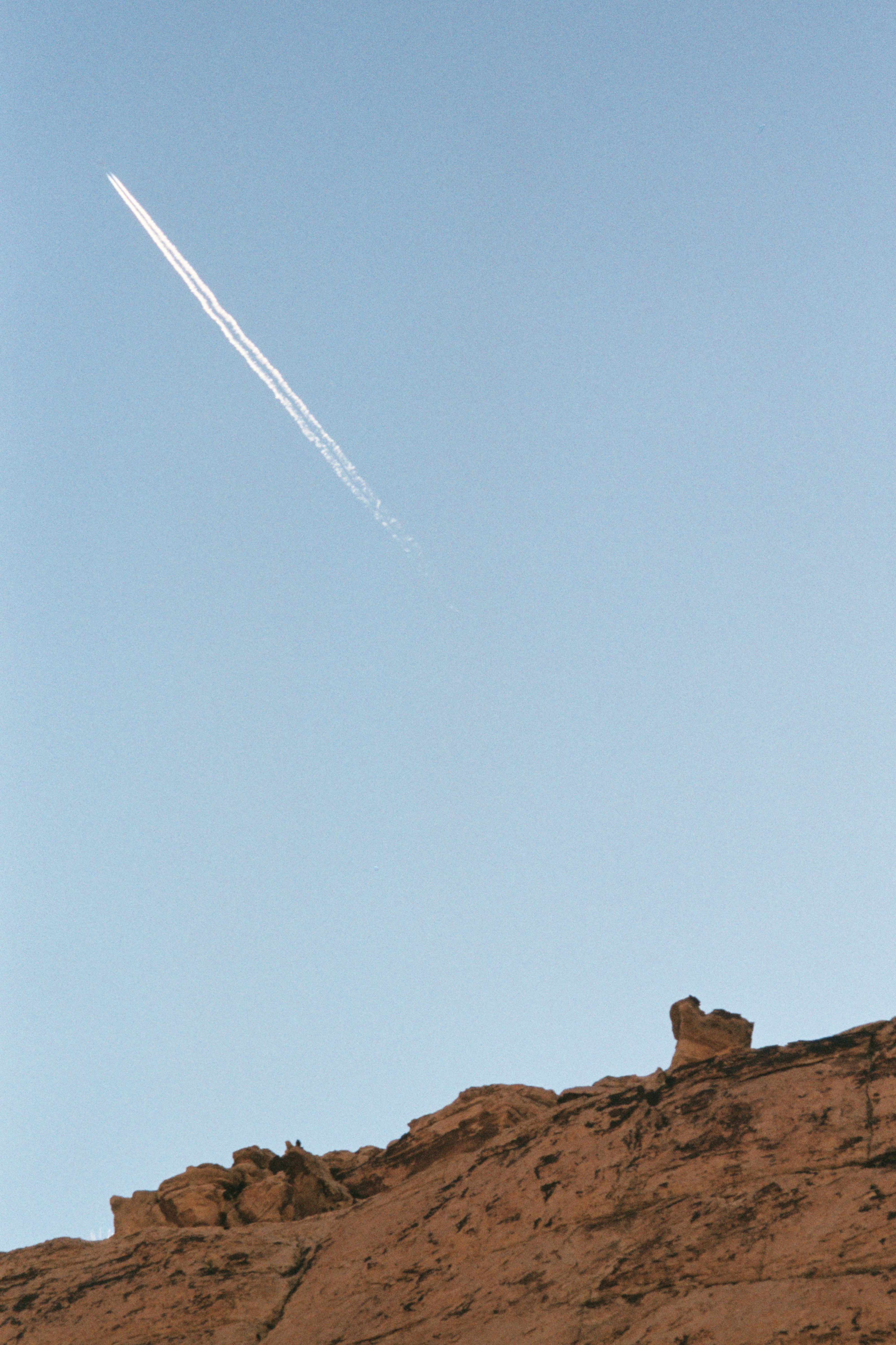 Contrail streaks across clear blue sky above rocky hillside.