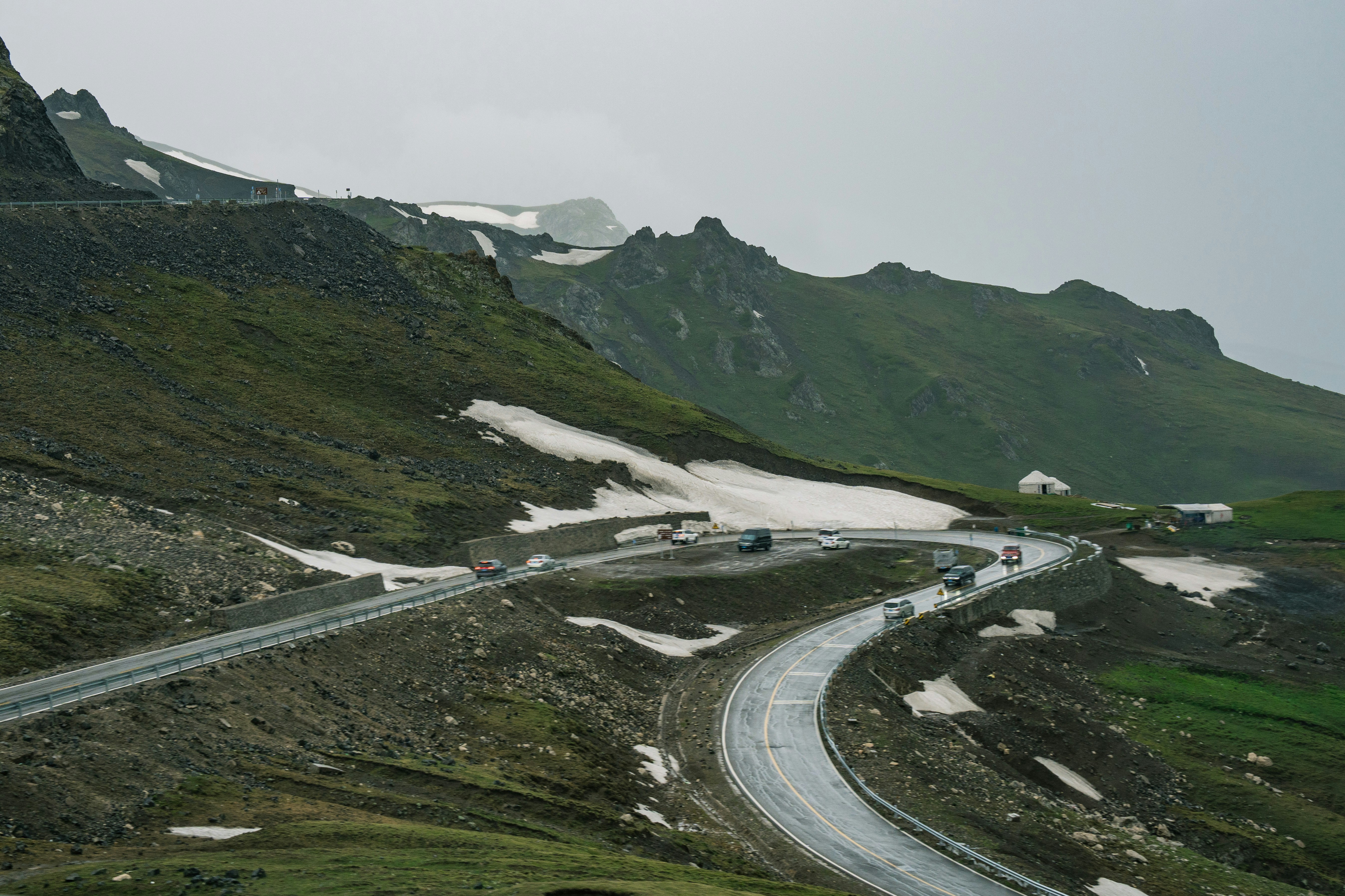 A winding mountain road with green slopes and snow patches.