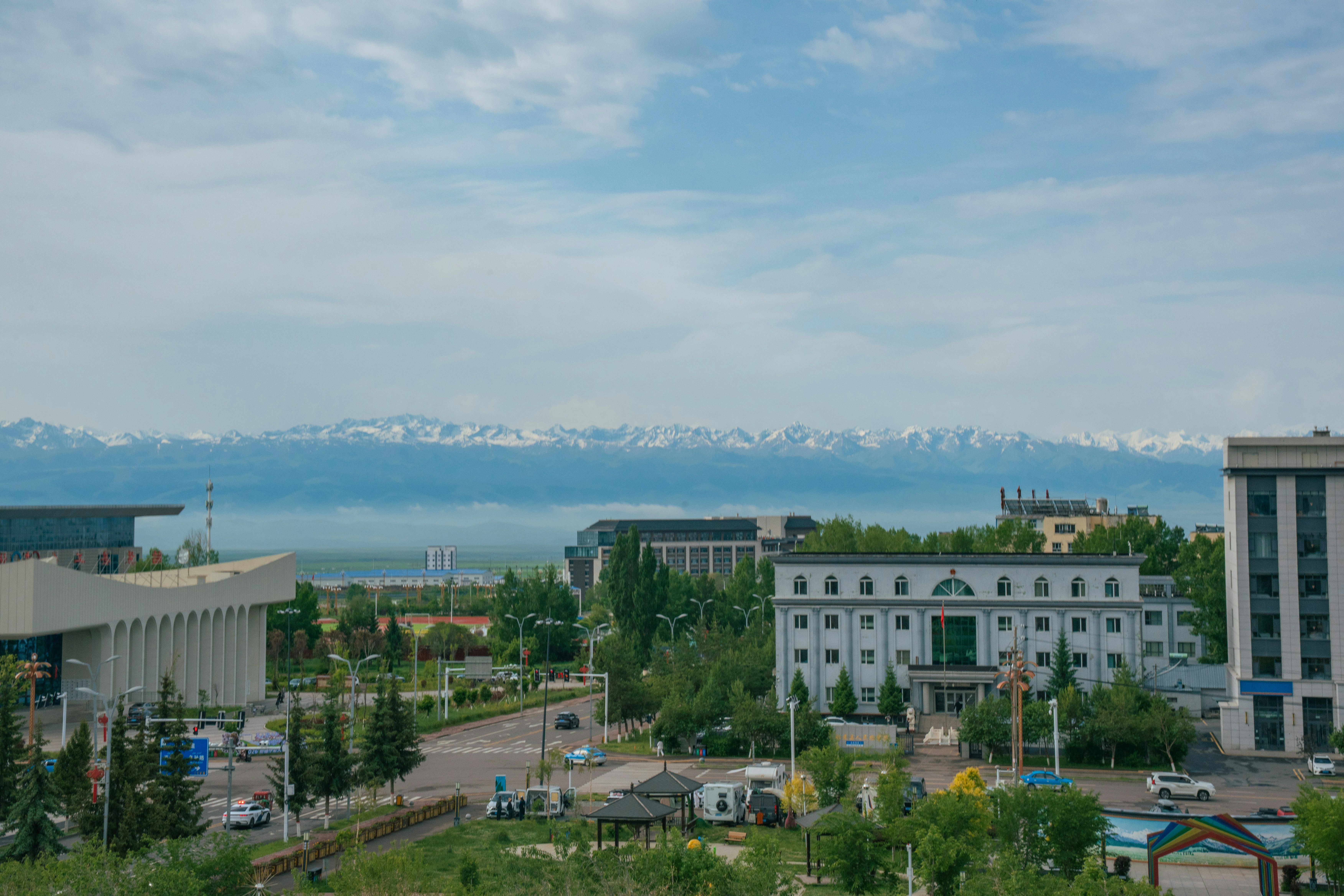 Cityscape with buildings, green trees, and distant snow-capped mountains.