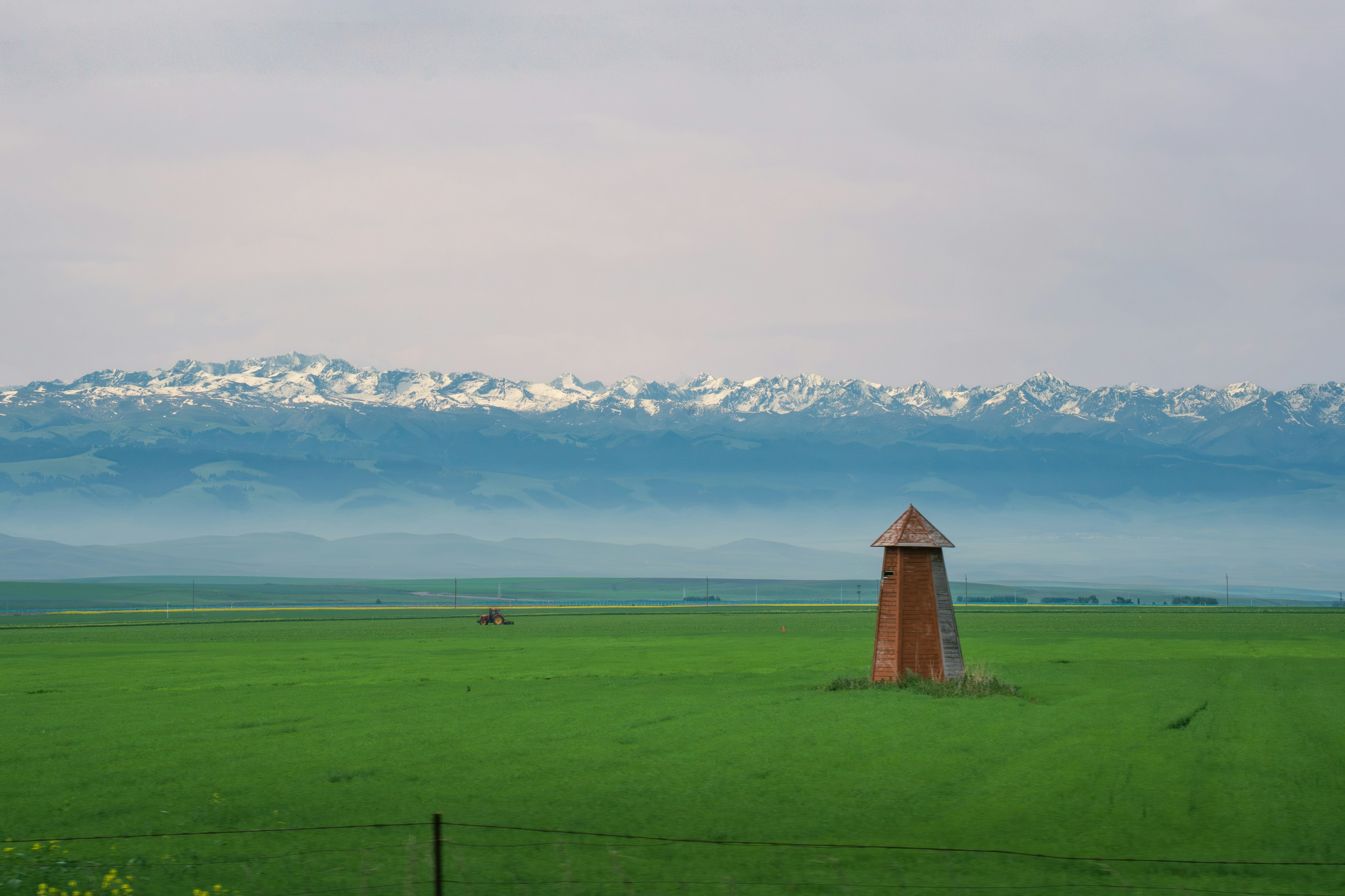 A wooden structure in a green field with snow-capped mountains.