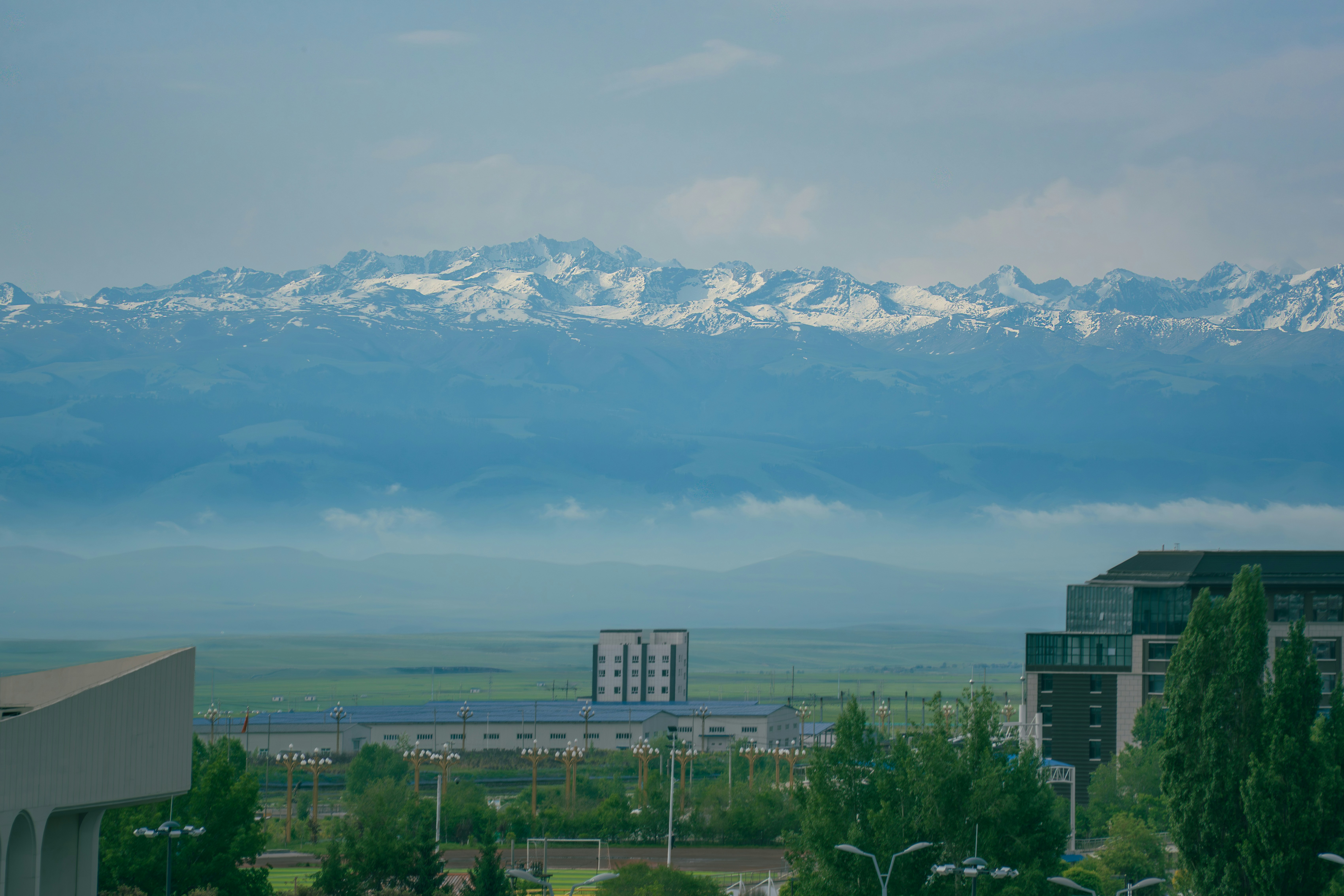Snow-capped mountains overlook buildings and a green landscape.