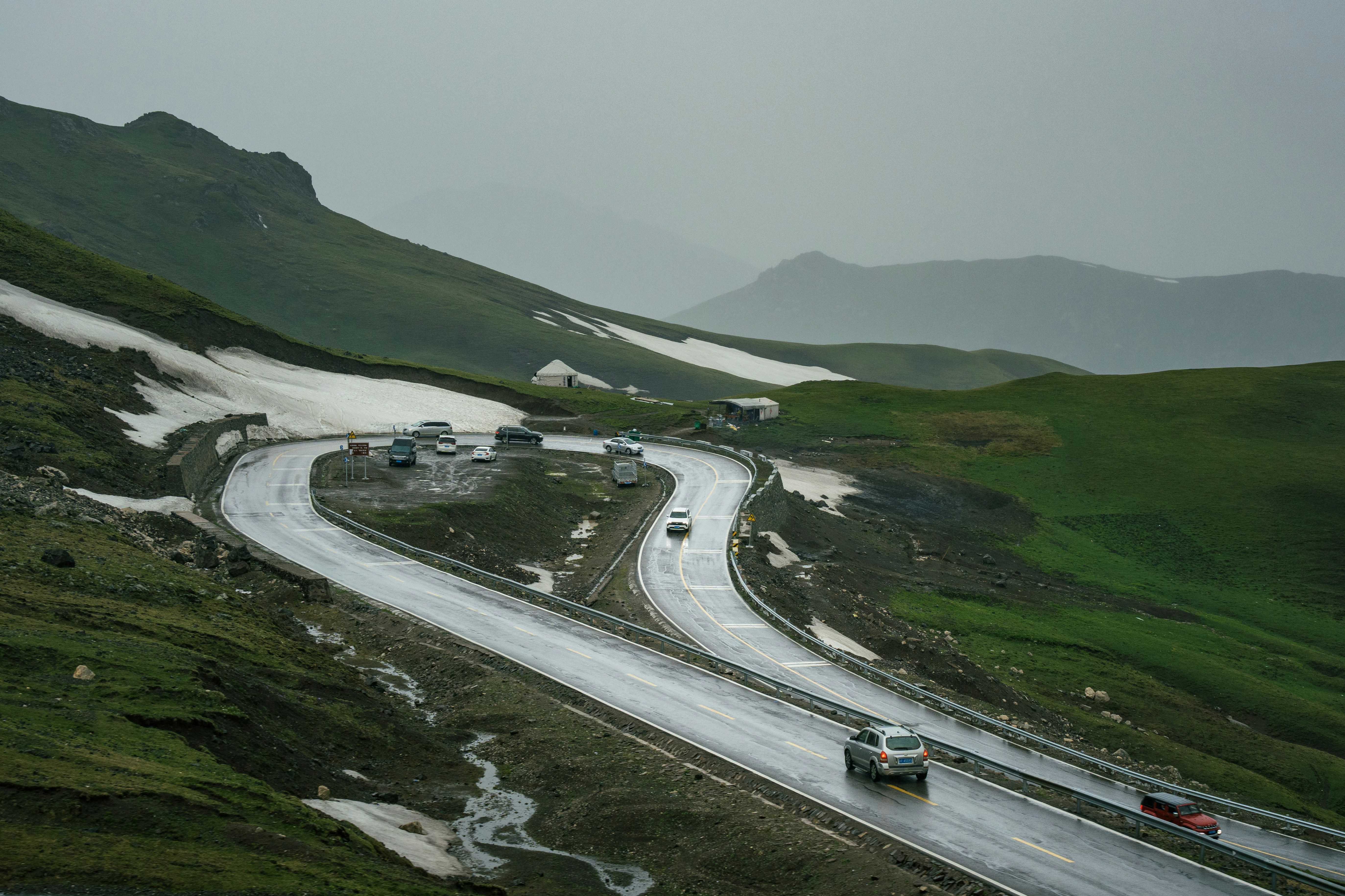 Rainy mountain pass with winding road, cars, and snow.