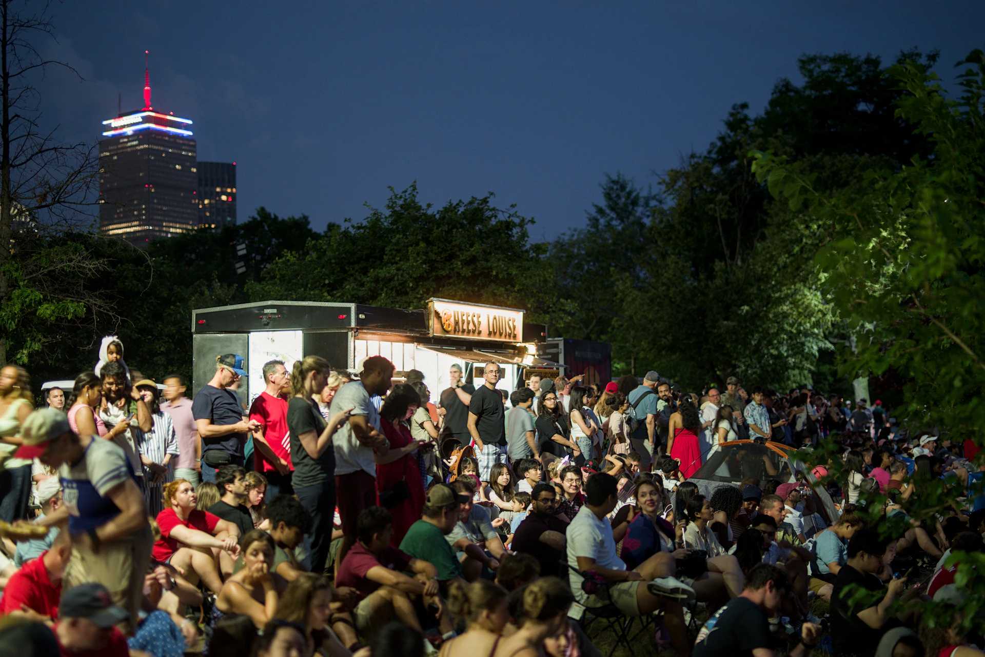 Crowd gathers outdoors at night, city skyline in background