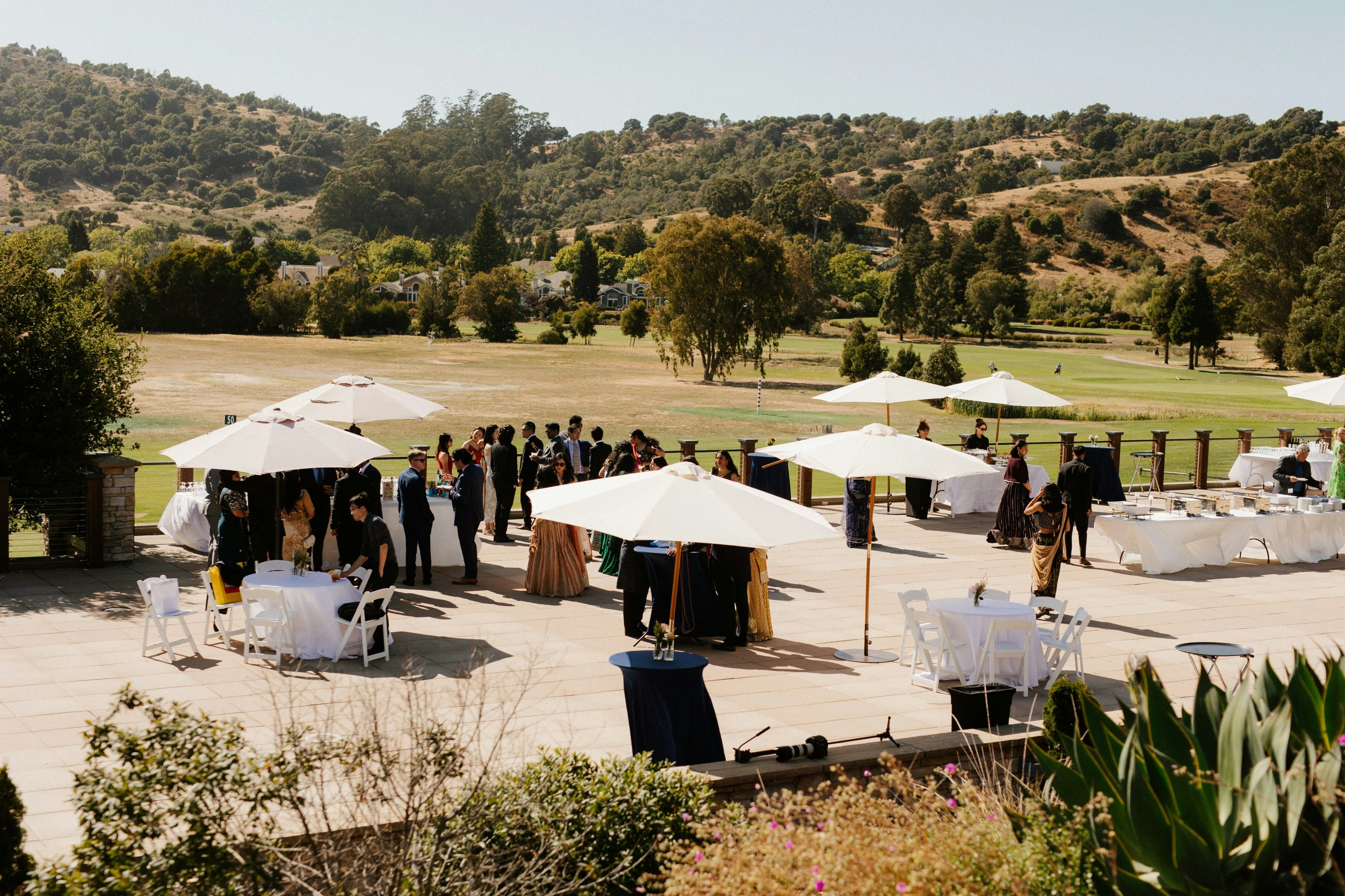 People gathered outdoors under umbrellas with hills in background.