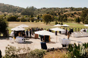 People gathered outdoors under umbrellas with hills in background.