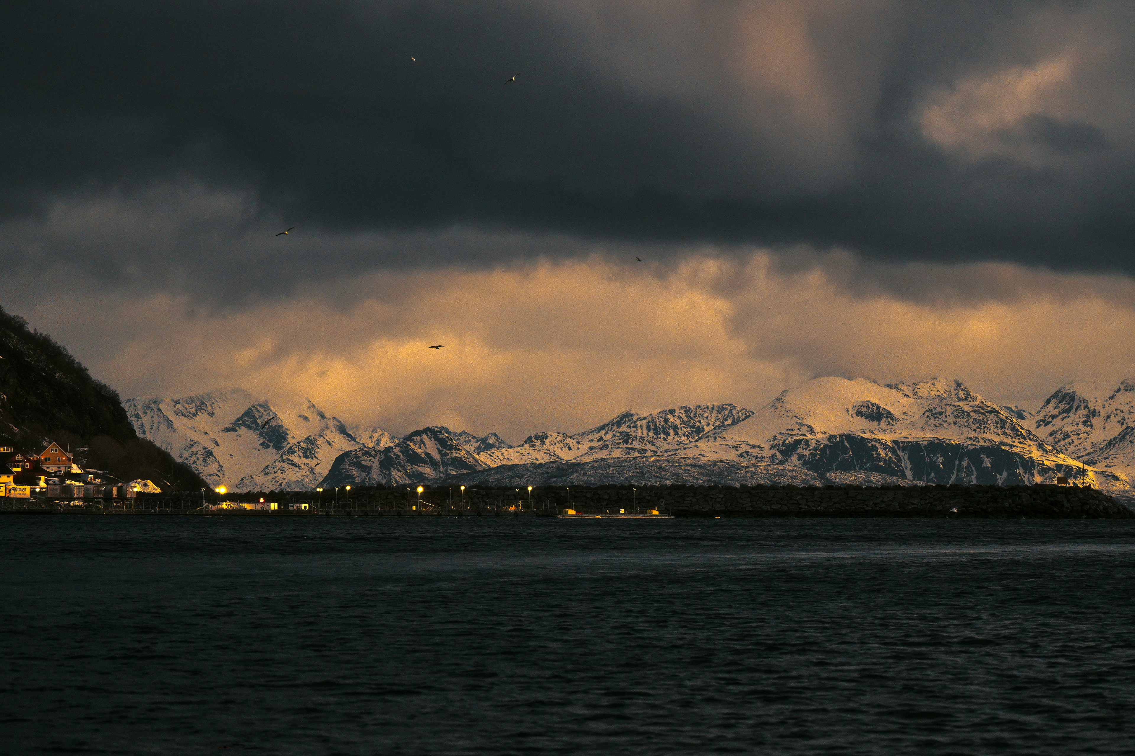 Dark clouds over snow mountains and a lit coastal village