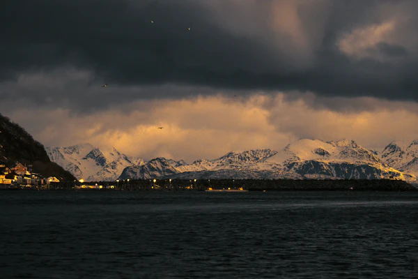 Dark clouds massed over snow-covered coastal mountains above a small lit village at dusk