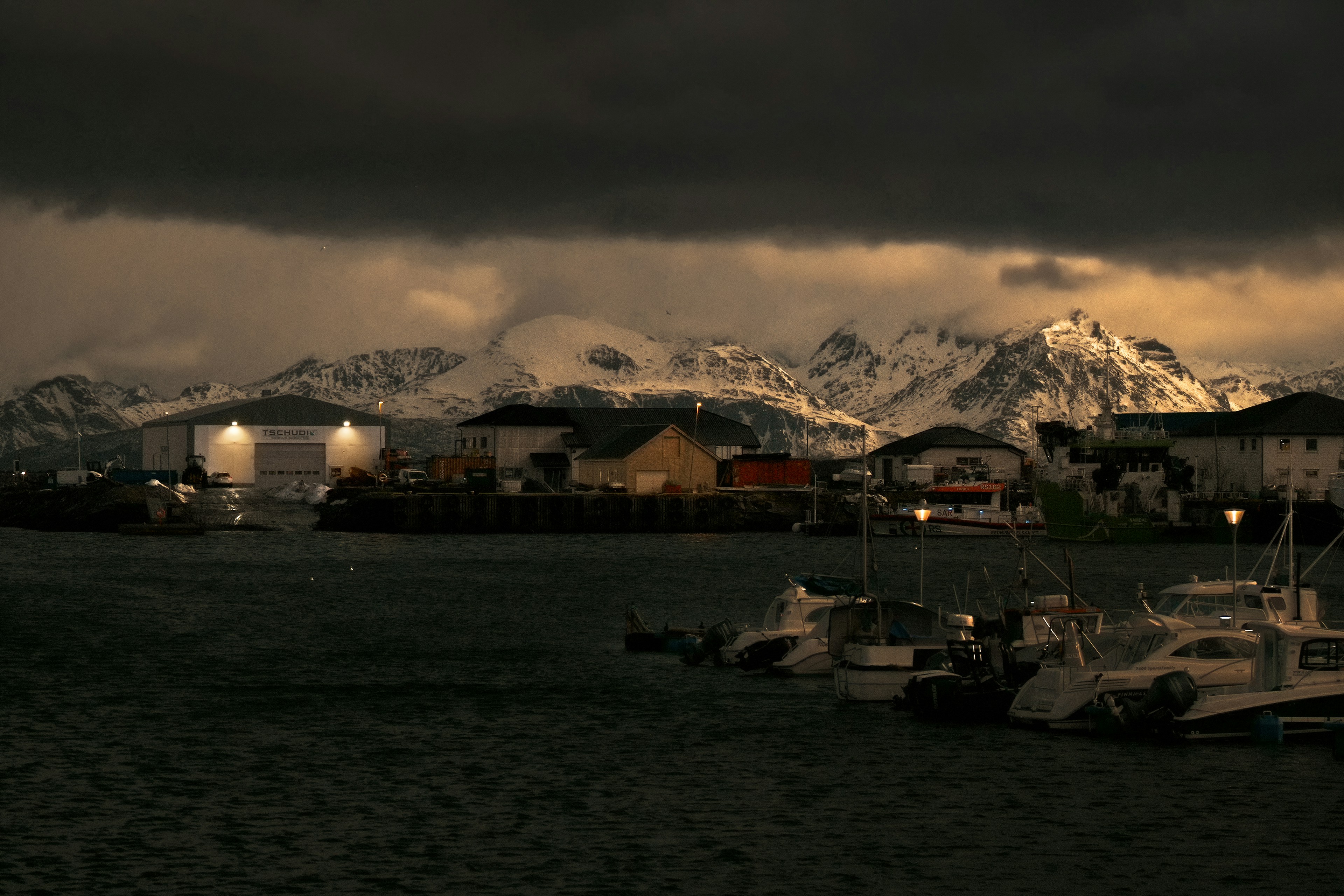 Dark clouds over a harbor with boats and snowy mountains.